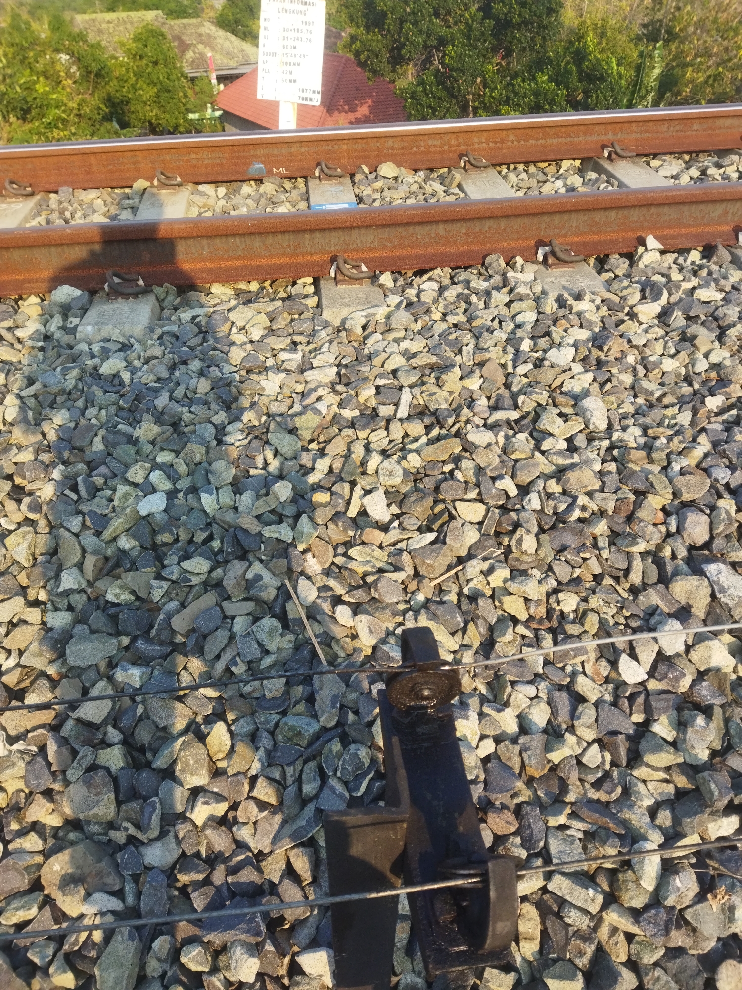 This is a medium-shot, eye-level, outdoor, daytime photograph of a section of train tracks with ballast. The tracks are made of rusty metal and are supported by concrete sleepers. The ballast consists of gray and tan stones. In the foreground, a black metal lever mechanism is visible, connected to thin wires. In the background, there are green trees and some buildings with red roofs. A white sign with text is also visible in the upper left quadrant of the image. This scene is located in Wonogiri, Indonesia, where train tracks are a common feature of the landscape, often found in proximity to residential areas and lush vegetation.