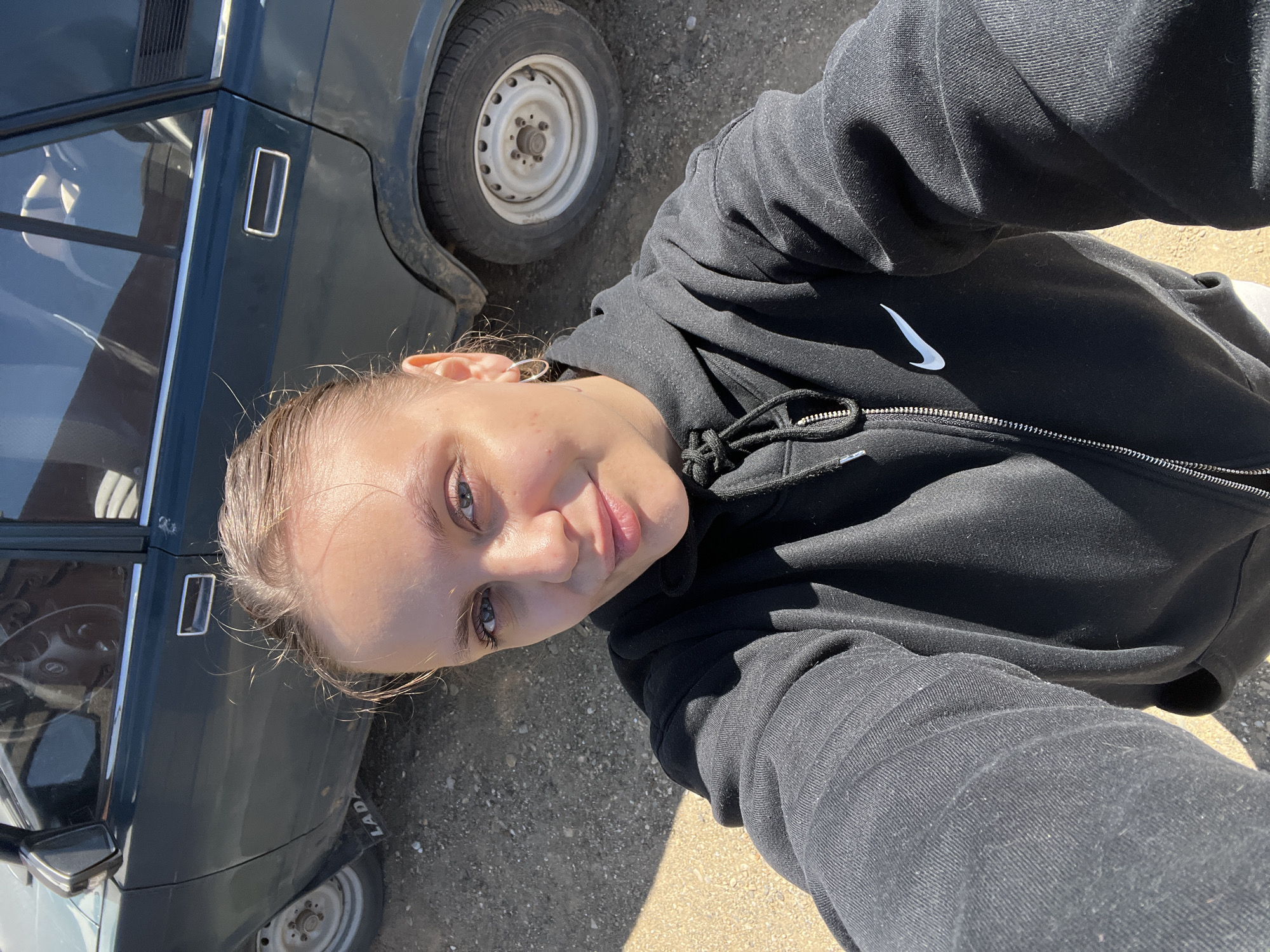 A young woman wearing a black Nike hooded sweatshirt takes a selfie next to an old dark green Lada car. The picture appears to be taken outdoors in a location with gravel or dirt ground. Based on the information provided and without any additional data, I can't verify whether this photo was taken in Ольховка, Russia.