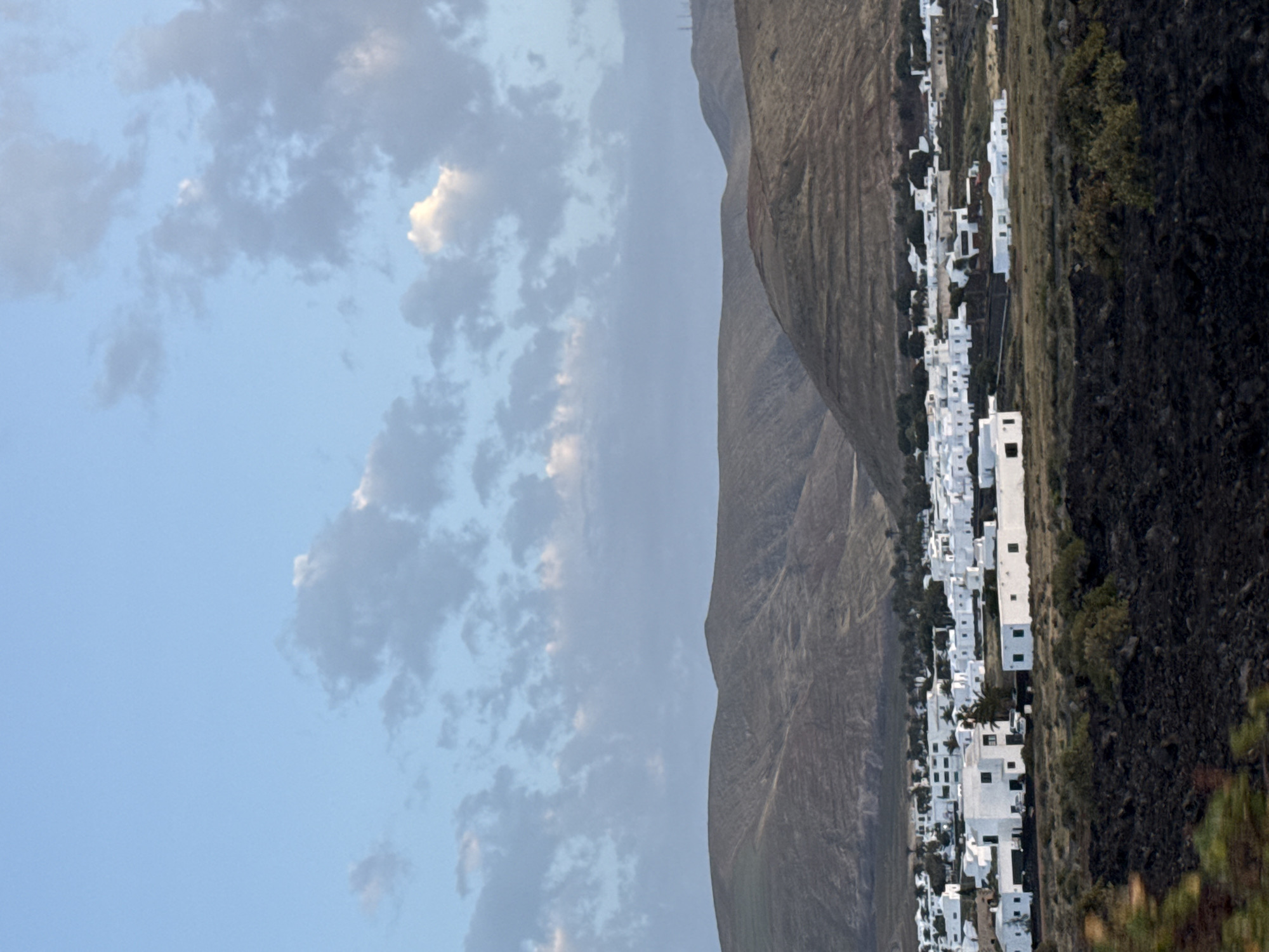The image depicts a hillside village in Yaiza, Spain, under a partly cloudy sky. A line of white, rectangular buildings snakes along the base of a mountain range. Adjacent to the buildings is a steep, black, rocky hillside with sparse vegetation. The sky is predominantly blue with scattered, light-gray clouds.