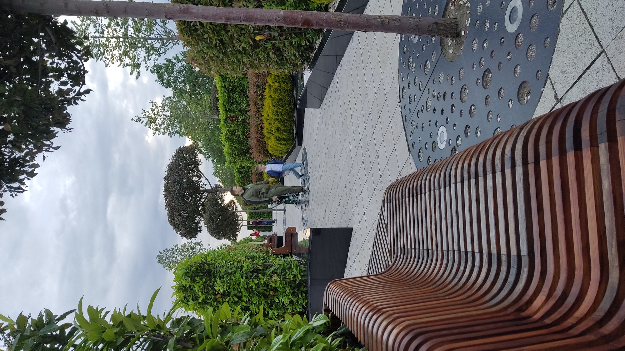 A vertically oriented photograph depicts a modern urban park or promenade in Krasnodar, Russia, under an overcast sky. The scene is characterized by contemporary landscaping and architectural elements.
In the lower right of the frame, a distinct wooden bench with numerous curved, dark brown slats forms a prominent foreground object. To its left, a dark circular metal tree grate, featuring various sized circular cutouts and inlaid with small stones or lights, encircles the base of a tree. The ground throughout the visible area is paved with light-colored square tiles.
Two main individuals are present in the mid-ground, standing on the paved path. One person, an adult dressed in olive-green attire, appears to be holding an item and faces a second, younger individual. This second person, possibly a child or teenager, wears a light blue denim jacket, light-colored pants, and white sneakers, and is looking towards the adult. Further in the background, several other people are partially visible near additional, simpler wooden benches along the pathway.
The setting is richly green, with a diverse array of plants and trees. Tall trees with reddish-brown trunks partially frame the left and right sides of the image. The path is bordered by dense bushes and hedges, showcasing various shades of green, yellow, and reddish foliage. Some trees in the background exhibit a sculpted or topiary form. A dark, low wall or planter runs along the edge of the path behind the main figures.
The weather appears overcast, with a cloudy sky providing soft, diffused light, indicating daytime without harsh shadows. The overall atmosphere suggests a calm and well-maintained public leisure space. No discernible text is visible within the image.