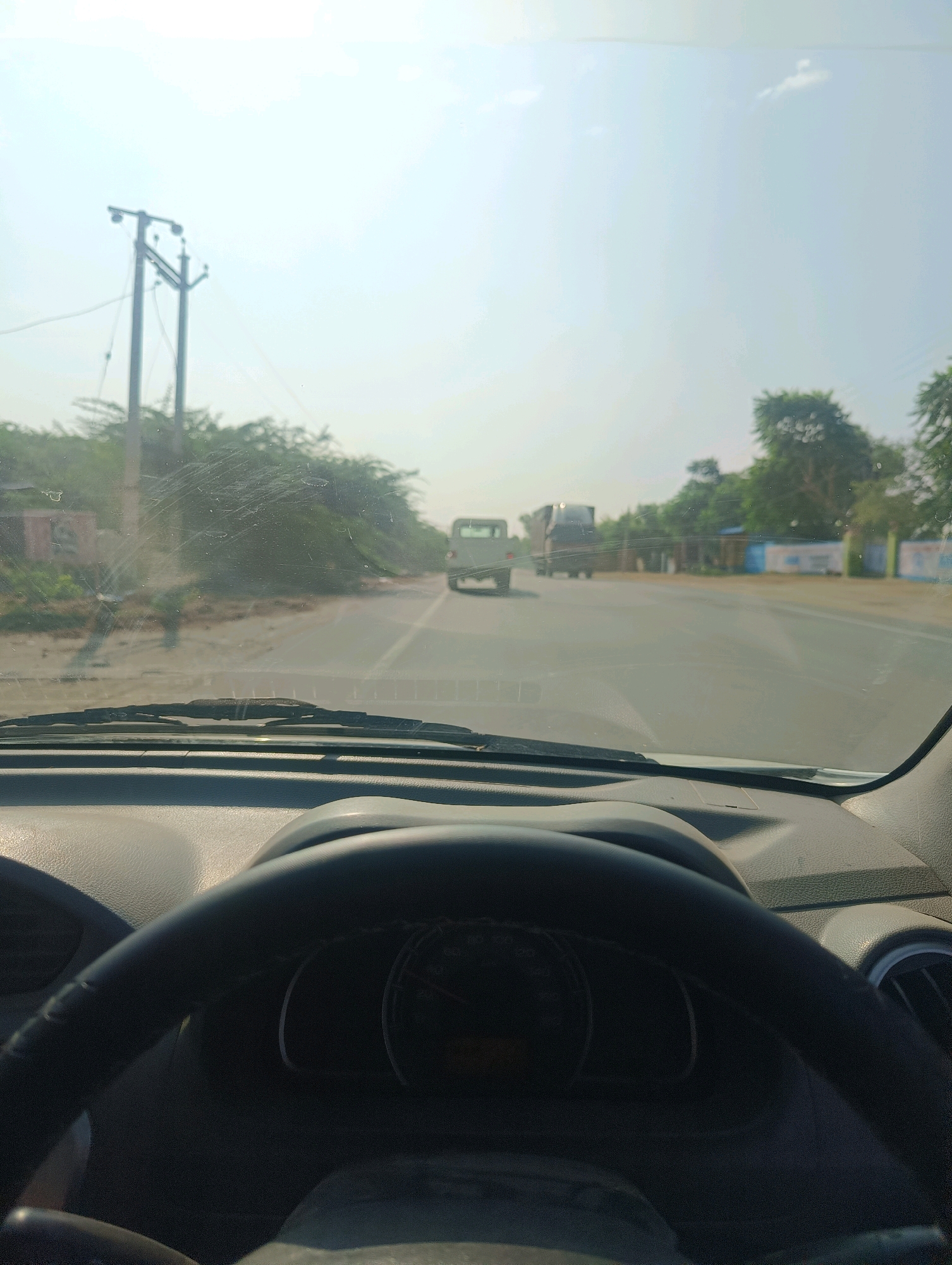 The image is taken from inside a car looking out through the windshield. The car is on a paved road, possibly a highway, with vegetation and trees lining the sides. In the distance, there are two trucks on the road. A utility pole with wires is visible on the left side of the road. The sky is clear and bright. The interior of the car is visible in the foreground, with a steering wheel and part of the dashboard. The scene appears to be in Chhapri Bari, India, given the context provided.