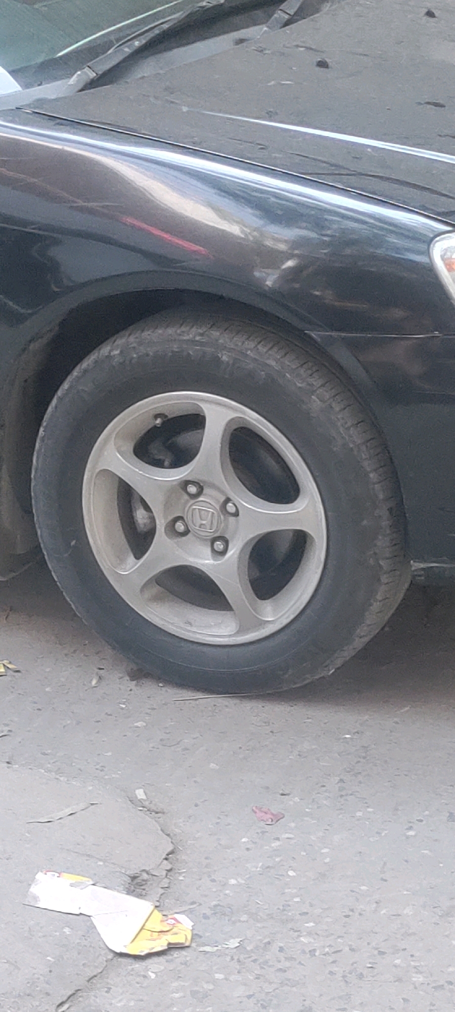 A close-up, low-angle shot focuses on the front passenger-side wheel and fender of a dark-colored car. The car's body is dusty, with reflections of light and possibly a red light source visible on the fender. The tire appears worn, and the silver alloy wheel has a five-spoke design with a central Honda logo. The wheel is positioned on a cracked, gray asphalt surface with scattered debris, including crumpled paper and a small pink object. The scene suggests an outdoor urban environment, likely a street or roadside within the Metropolitan Corporation Rawalpindi, Pakistan, due to the characteristic road surface and common vehicle type.