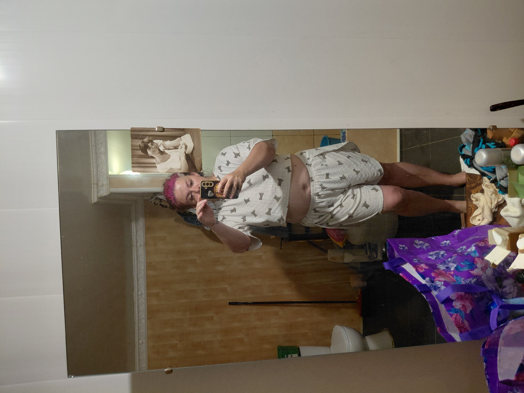 A full-body mirror selfie is taken in a bathroom, with the photographer holding a phone to capture the image. The photographer, who appears to be a woman with pink hair, is wearing a light-colored crop top and shorts with a repeating pattern of small gray cats. In the reflection of the mirror, a toilet and a broom are visible in the background. Above the reflection of the bathroom, a framed black and white photograph of a woman on the phone is attached to the wall. The bathroom walls are covered in a wood-patterned wallpaper. There are several bags and items on the floor in front of the mirror, including a large purple floral bag. The overall environment appears to be a residential bathroom. The location is identified as Marynivka, Ukraine.