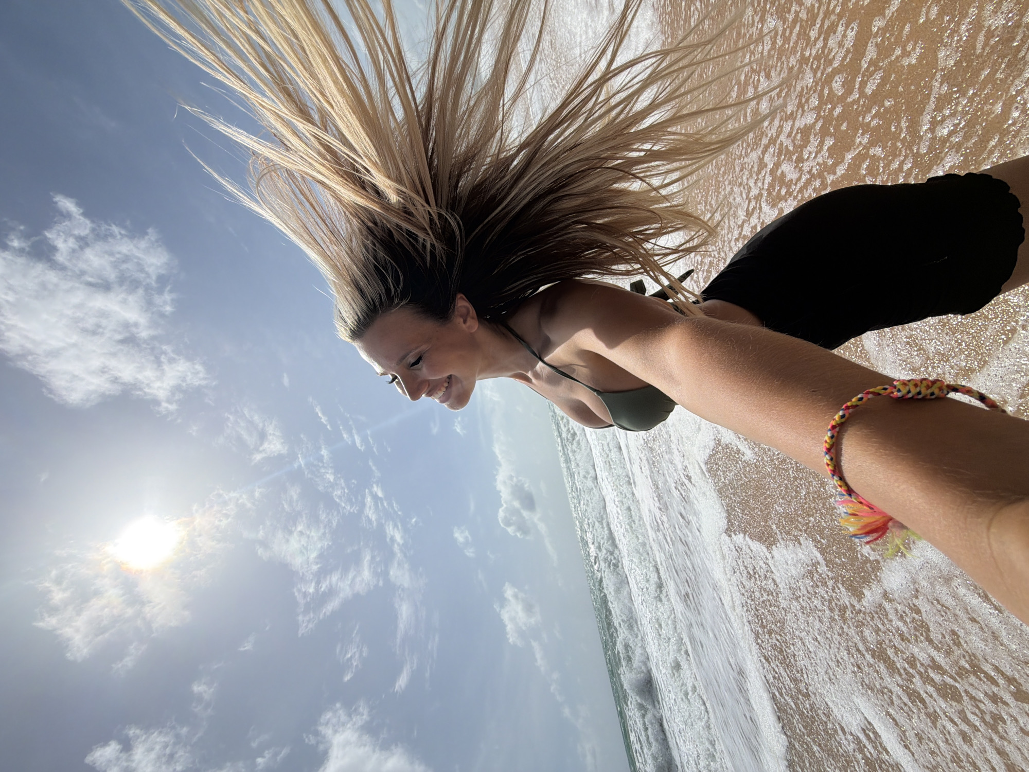 A young woman with long, blonde hair blowing in the wind stands on a beach in Mai Khao, Thailand, as waves crash onto the shore. She is wearing a green bikini top and black shorts, and a colorful bracelet adorns her wrist. The sun is high in the sky, casting a bright glare through the clouds. The scene is captured from a low angle, with the woman's arm extended forward, suggesting she is taking a selfie. The water is a deep blue-green, and the sand is a light brown.