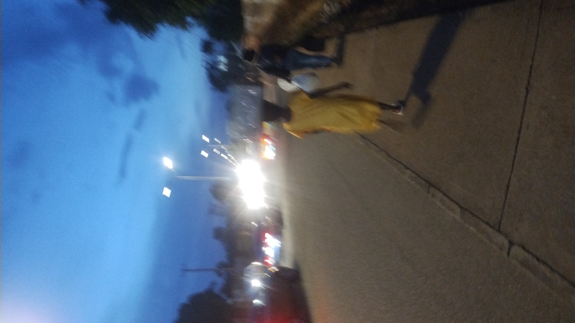 The image depicts an outdoor street scene at dusk or night, characterized by strong artificial lighting and a dark blue sky. The perspective is rotated, showing the sky on the left and a street and sidewalk on the right.
On the left side, the sky is a deep blue, with faint, darker cloud formations visible. Several bright, square streetlights are mounted on poles, illuminating the scene.
On the right side of the image, a sidewalk runs parallel to a road. A prominent figure is walking or standing on the sidewalk, wearing a bright yellow shirt or dress. A long, dark shadow extends behind this person, cast by an unseen light source. Further down the sidewalk, other blurred figures are visible, some in darker clothing like jeans.
The adjacent road is occupied by vehicles. Intense glare from bright white headlights dominates the mid-ground, making it difficult to discern specific vehicle details. Red taillights from other vehicles are faintly visible further back along the street. The overall impression is one of movement, with people and vehicles active in a low-light urban environment.
The photograph is somewhat blurry and exhibits motion blur, particularly around the lights and moving figures, suggesting a low-light capture or a slow shutter speed. No text is visible in the image.