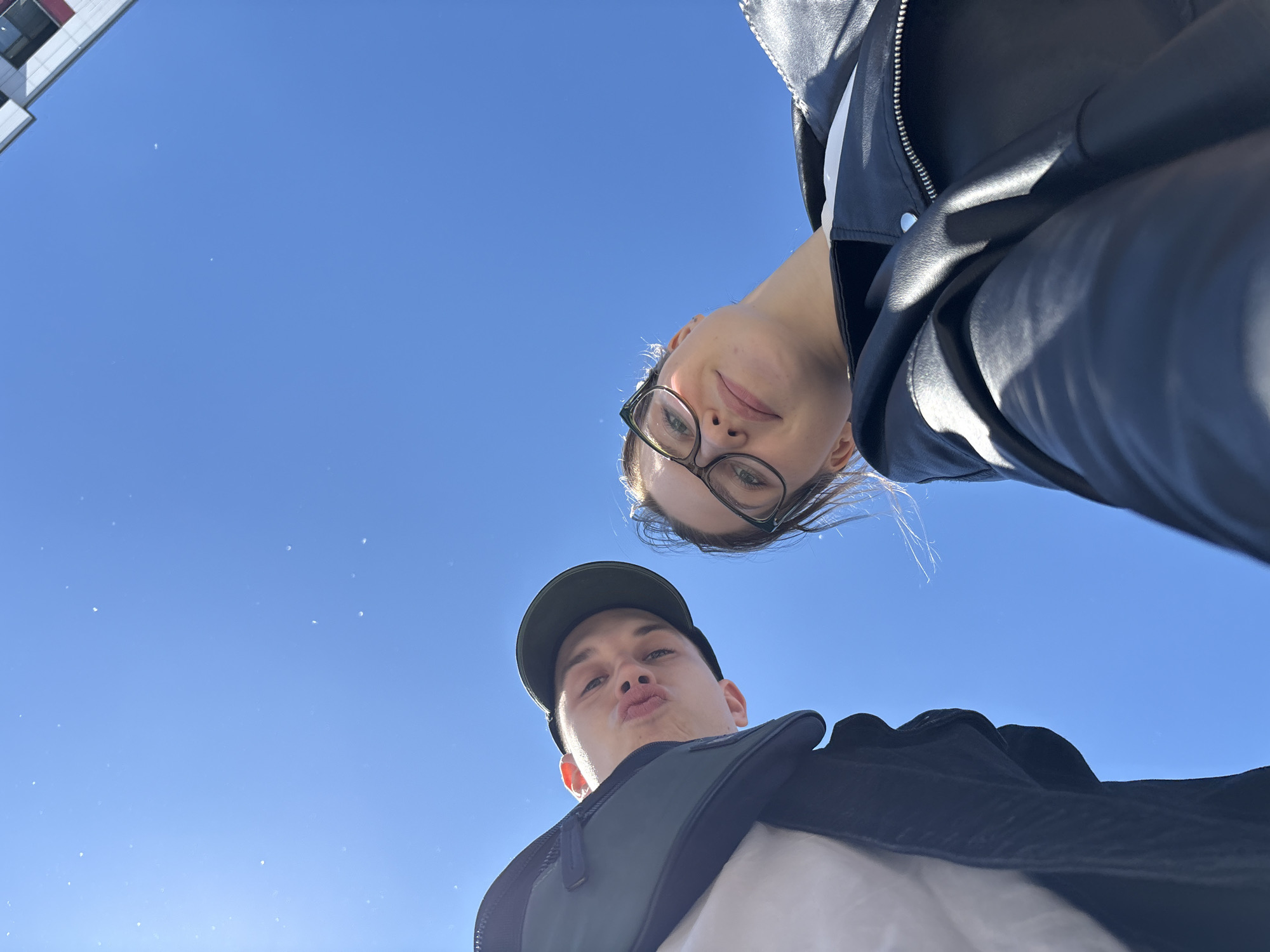 This is a low-angle, wide shot selfie taken outdoors on a clear, sunny day. Two young adults, a woman and a man, are prominently featured, looking up at the camera. The woman, on the left, wears glasses and a black leather jacket over a white garment. Her face is tilted downwards, and she has a slight smile. The man, on the right, is wearing a dark baseball cap and a black jacket or vest over a white shirt. He is puckering his lips as if for a kiss and looking directly at the camera. The background is a vibrant, cloudless blue sky with a few small, indistinct white specks, possibly dust or lens flare. In the upper left corner, a portion of a modern building with a white and red facade is visible, suggesting an urban or semi-urban environment. The lighting indicates it is daytime, likely midday due to the bright sun and clear sky. The overall mood appears playful and casual. No visible text is present in the image. The scene evokes a sense of youthful spontaneity and enjoying a bright day, with the Saint Petersburg, Russia location context suggesting a potential backdrop of the city's architecture.