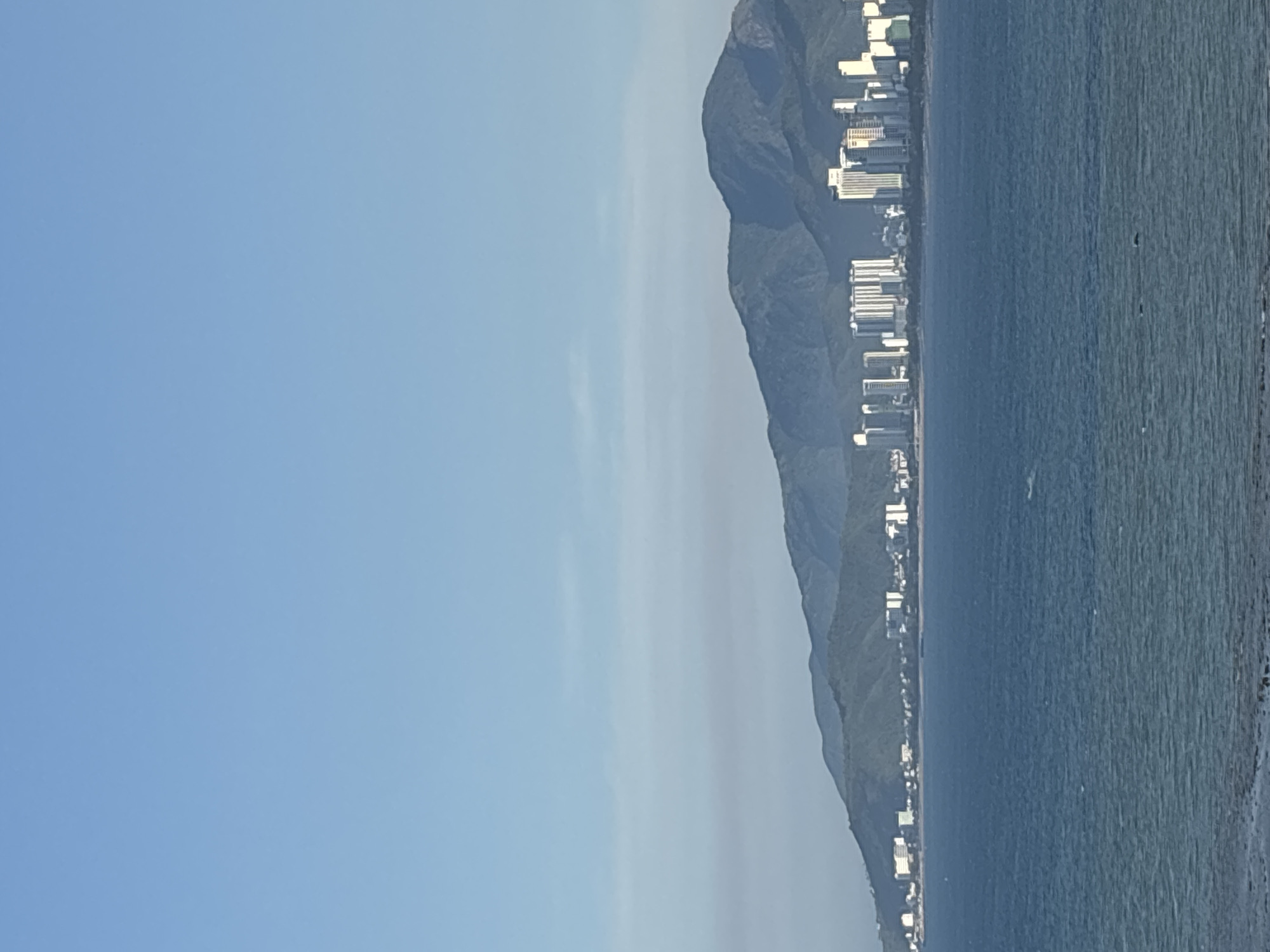 A wide, horizontal shot shows a cityscape nestled against a backdrop of verdant, mountainous terrain, all under a clear, cerulean sky. In the foreground, a deep blue ocean stretches across the lower third of the frame, its surface rippled with gentle waves. A faint, rocky shoreline is visible at the very bottom.
Dominating the mid-ground is a densely packed urban skyline, characterized by numerous high-rise buildings of varying heights and architectural styles. The buildings are predominantly white, with some featuring darker or colored accents. They appear to be modern structures, likely hotels or residential complexes. The cityscape lines the coast, separating the ocean from the imposing mountain range behind it.
The mountains themselves are covered in lush, green vegetation, with multiple peaks and valleys creating a dramatic natural silhouette against the sky. The lighting suggests it is daytime, possibly morning or late afternoon, given the soft light and the absence of harsh shadows. The overall impression is one of a coastal city with a strong natural landscape.