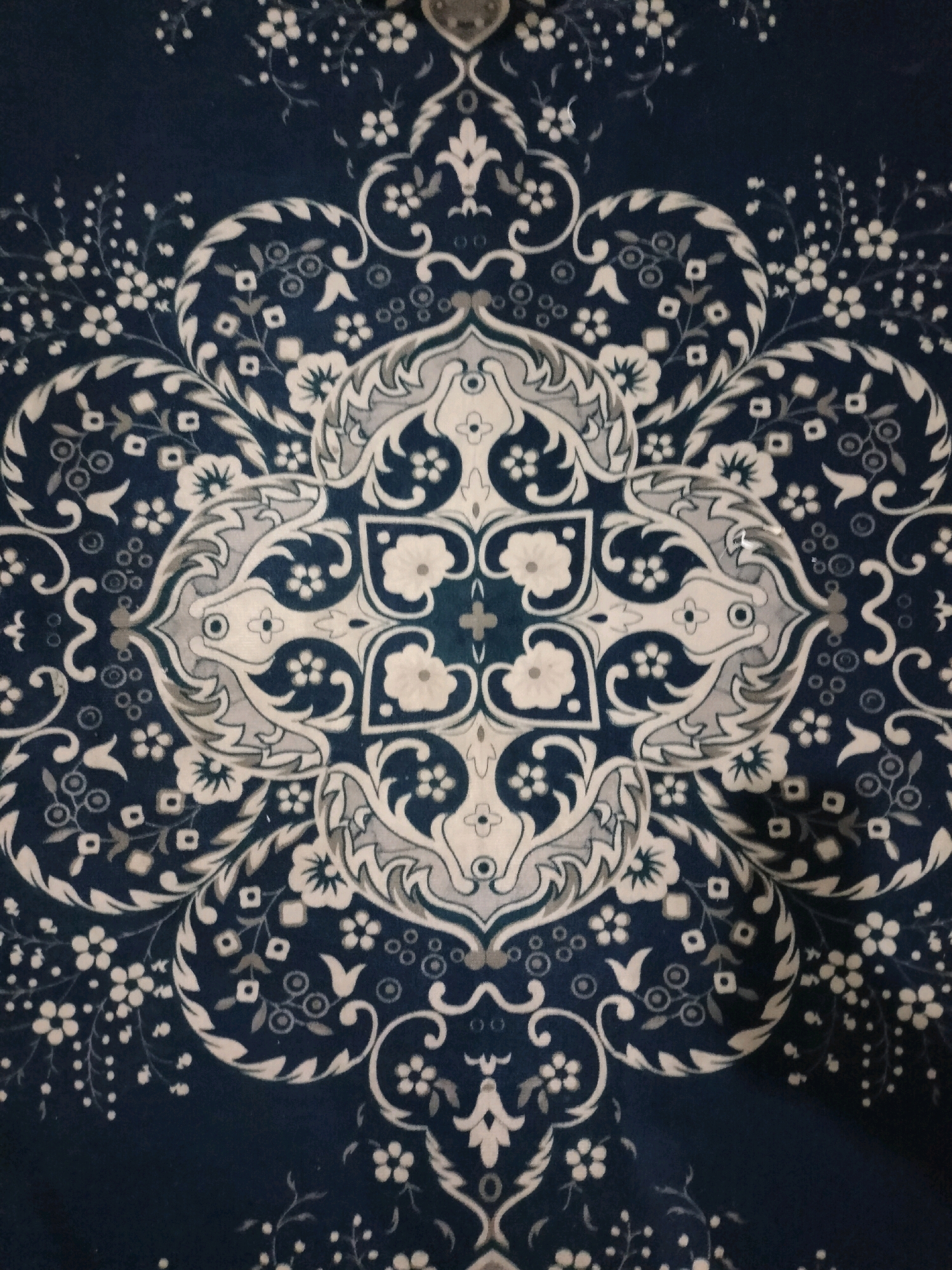 The image is a close-up, top-down view of a dark blue fabric with a white and gray ornamental pattern. The pattern is symmetrical and features a central, intricate design with floral and paisley-like elements radiating outwards. Scattered throughout the fabric are smaller floral motifs and dot-like elements. The lighting suggests a dimly lit environment, possibly indoors, with a slight shadow cast on the lower right side of the fabric. There are no people, actions, or interactions visible in the image. The location in Wedung, Indonesia, is not visually discernible from the provided image.