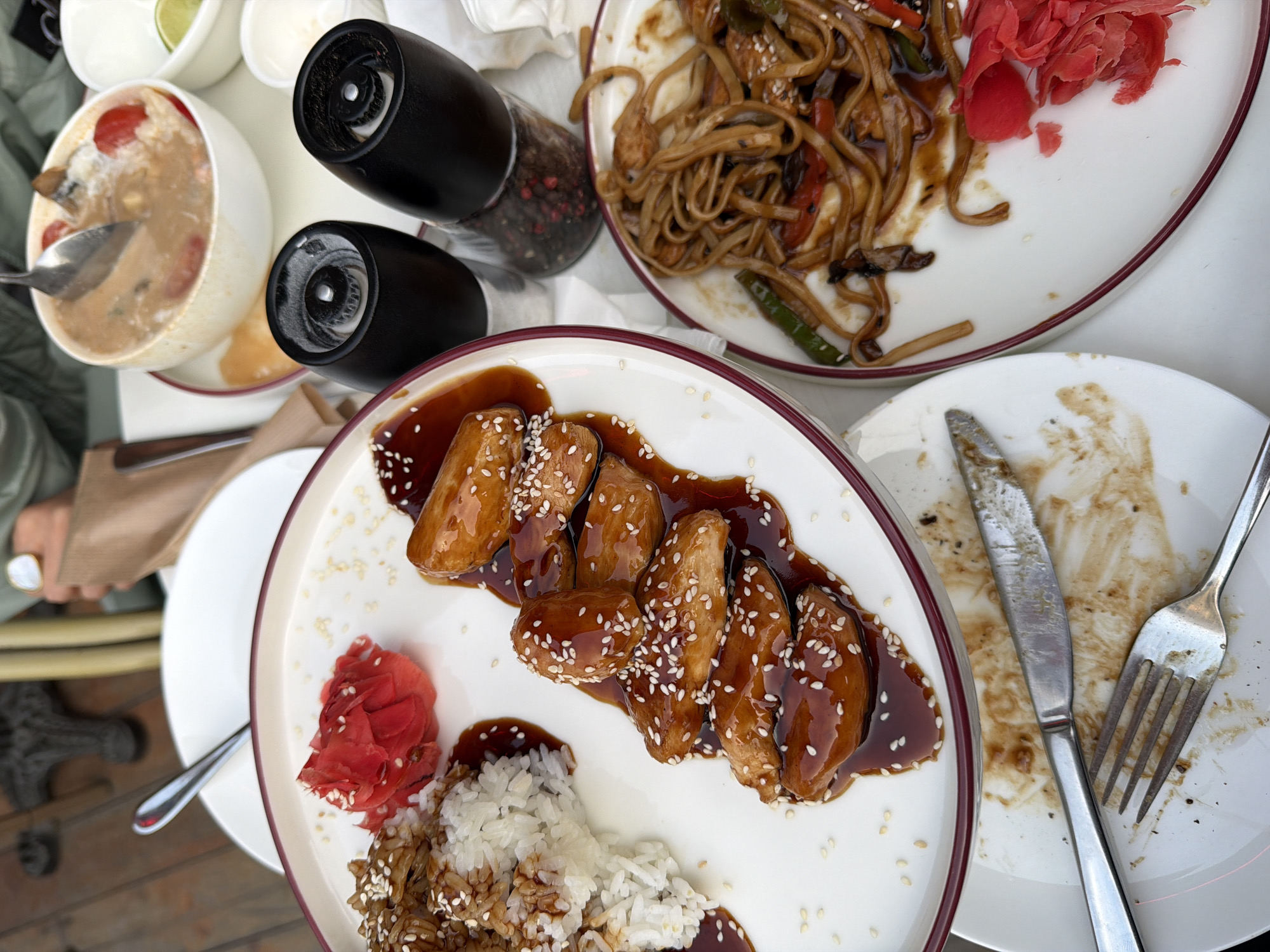 The image presents a table setting with several dishes, likely in Yerevan, Armenia, given the provided information. A white bowl containing soup with visible tomatoes sits to the left, accompanied by two black pepper grinders. A plate of noodles with vegetables and red ginger is placed nearby. In the foreground, a white plate with a red rim holds pieces of food covered in a dark sauce and sprinkled with sesame seeds, alongside rice and red ginger. An adjacent plate shows signs of being finished, with a knife and fork resting on it.