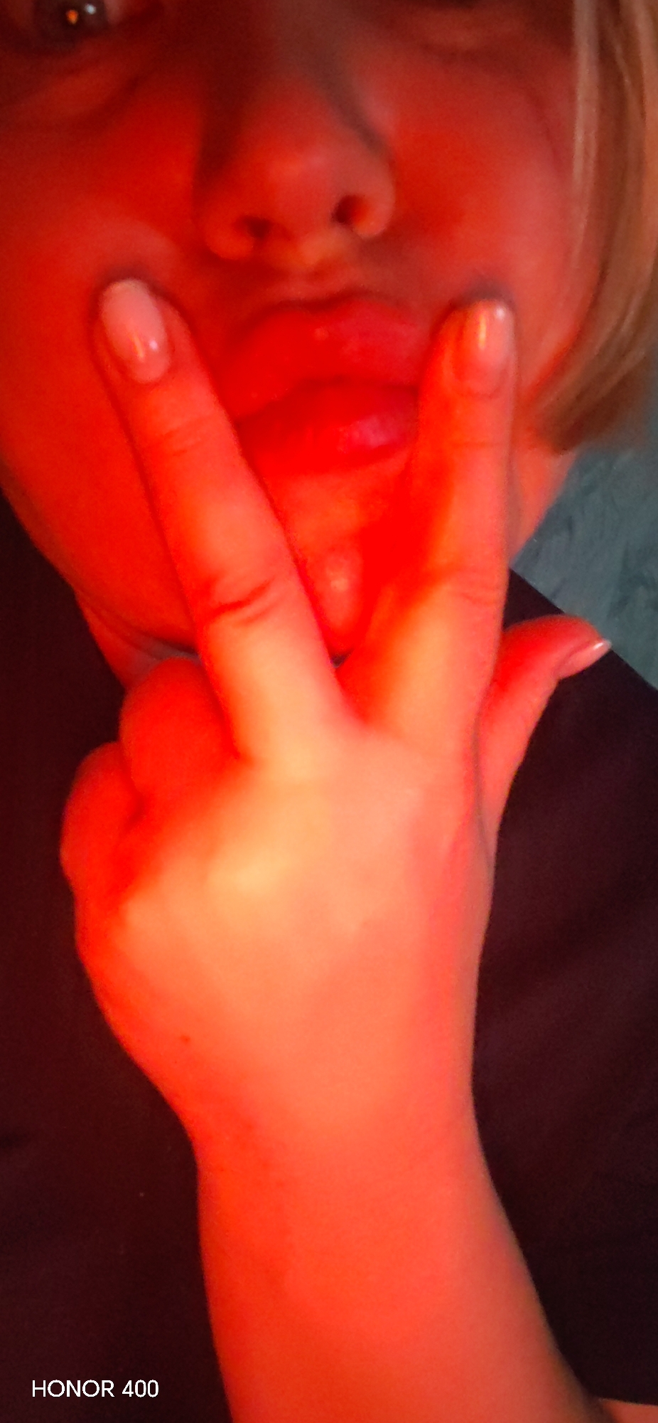 The image displays a close-up, vertically oriented portrait of an individual illuminated predominantly by a strong red light. The upper portion of the subject's face is visible, including a partially visible left eye, the nose, and the mouth. The lips are pursed, suggesting a specific facial expression. A left hand is positioned prominently in the foreground, performing a gestural sign where the index finger and pinky finger are extended upwards, while the middle and ring fingers are folded down towards the palm. The extended fingernails have a light, opaque polish. The individual appears to be wearing dark-colored clothing, possibly black, visible on the shoulders and chest. The background is largely obscured by darkness and the intense red lighting, with a faint lighter area, possibly a blue or teal surface, visible behind the subject's right shoulder. There are no discernible environmental cues to indicate a specific location in the city of Noviy, Russia, or any other location. The text "HONOR 400" is visible in the bottom left corner, indicating the imaging device.