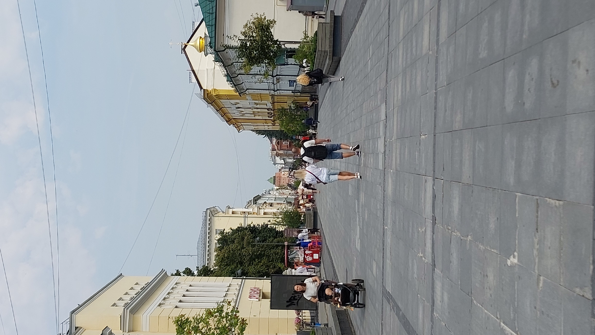 This image depicts a pedestrian street in Nizhny Novgorod, Russia. The street is paved with large gray tiles and is lined with historical buildings on both sides. The buildings feature a mix of architectural styles, with some exhibiting classical elements and others showcasing more colorful facades.
In the foreground, a woman is pushing a stroller with a baby. Further down the street, a couple is walking away from the camera, and other pedestrians are visible in the distance. Several food stalls and kiosks are set up along the sides of the street, indicating a lively commercial area. Trees and greenery are planted in planters along the street, adding to the urban landscape. The sky is overcast with a few scattered clouds. In the far distance, a tower is visible, which appears to be part of the Nizhny Novgorod Kremlin. The overall impression is of a pleasant, bustling urban environment on a slightly cloudy day.