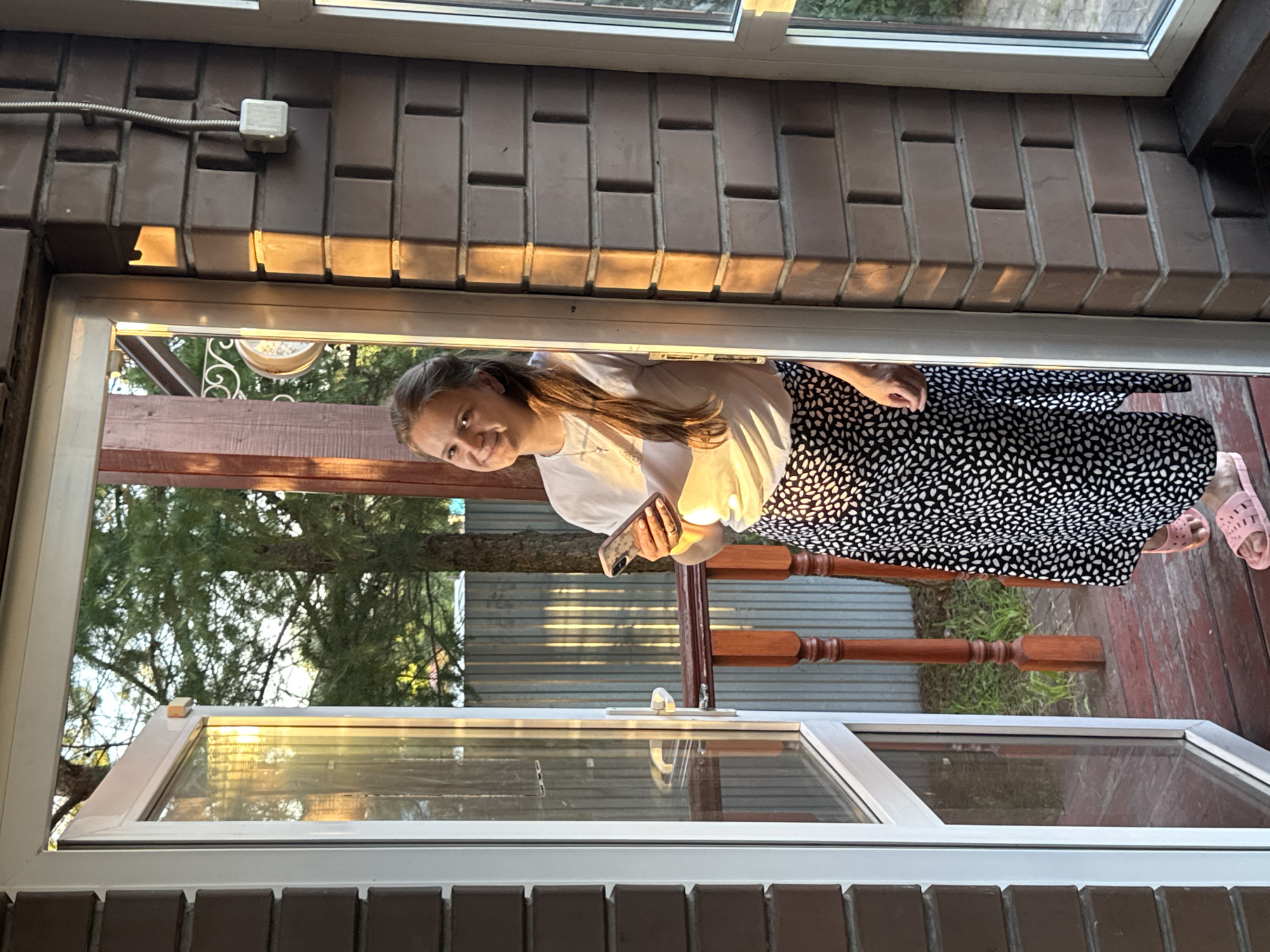 A young woman with long brown hair is standing in an open doorway, holding a smartphone. She is wearing a white t-shirt and a black and white patterned skirt. She is smiling and looking at the camera. The doorway opens onto a wooden porch, with a wooden railing and a metal fence in the background. There are trees and sunlight visible outside. The woman is wearing pink slide sandals. The wall next to the doorway is made of brick. The image was taken in Perm, Russia.