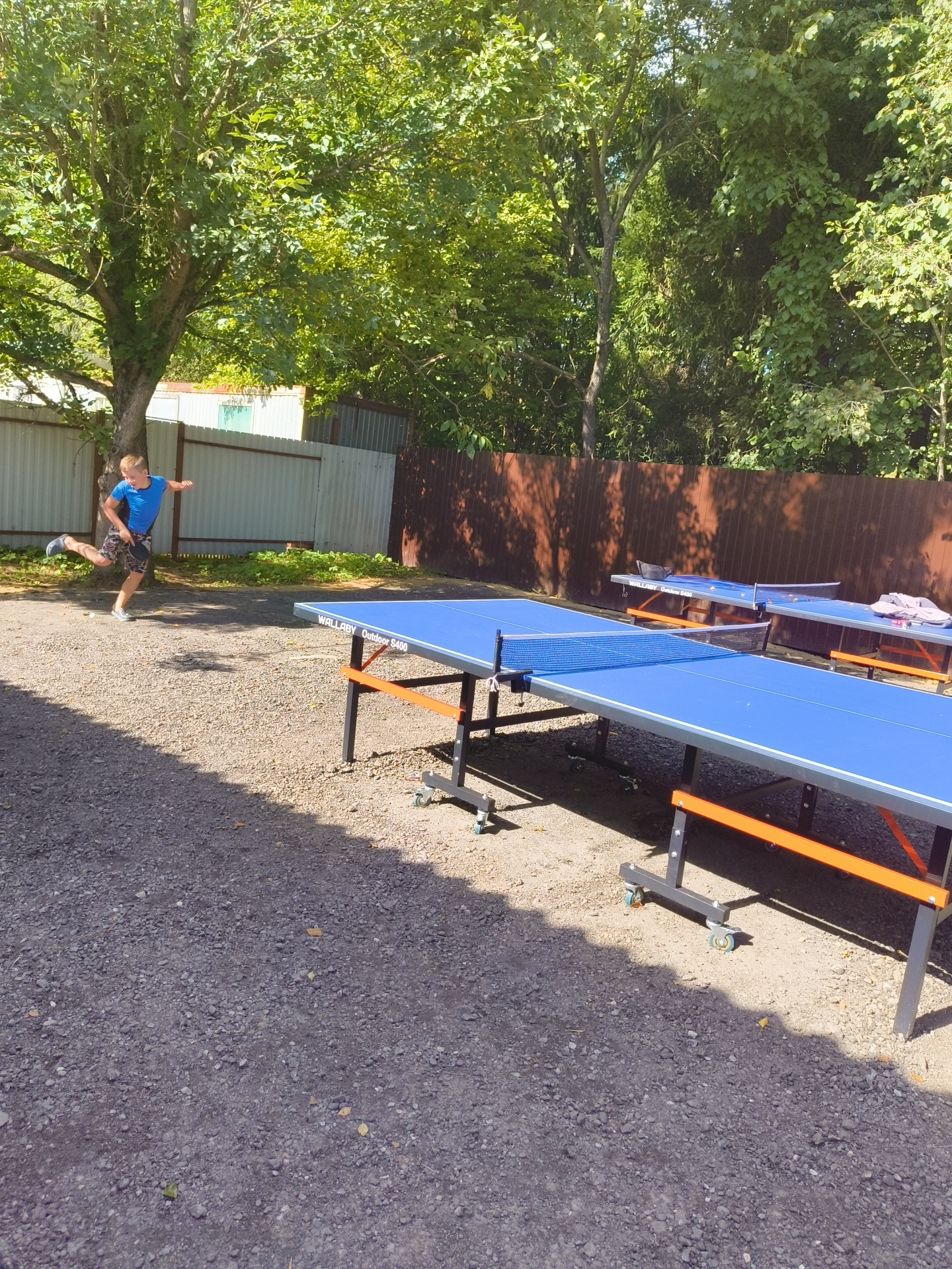 In the city of Pravdist-2 SNT, Russia, on a sunny day, a young Caucasian boy with blonde hair, wearing a blue t-shirt and patterned shorts, is captured mid-action, playing ping pong outdoors. He is positioned in the left half of the frame, leaning forward with his right leg extended and his left arm raised, holding a ping pong paddle. His gaze is directed towards something out of frame to his right, suggesting he is about to hit a ball or has just done so.
The scene is set on a gravelly surface, with two bright blue ping pong tables dominating the foreground and mid-ground. The table on the right is closer to the viewer and displays a net. The table further back is partially obscured by the first, and another table is visible behind that, suggesting a recreational area. The tables have dark metal frames with orange accents.
Behind the tables, a brown wooden fence runs horizontally across the mid-ground, separating the playing area from a background filled with lush green trees and foliage, indicating a natural, possibly park-like setting. In the far left background, a white structure with some green elements is visible behind the fence, possibly a building or shed. The lighting suggests it is daytime, with strong sunlight casting shadows.