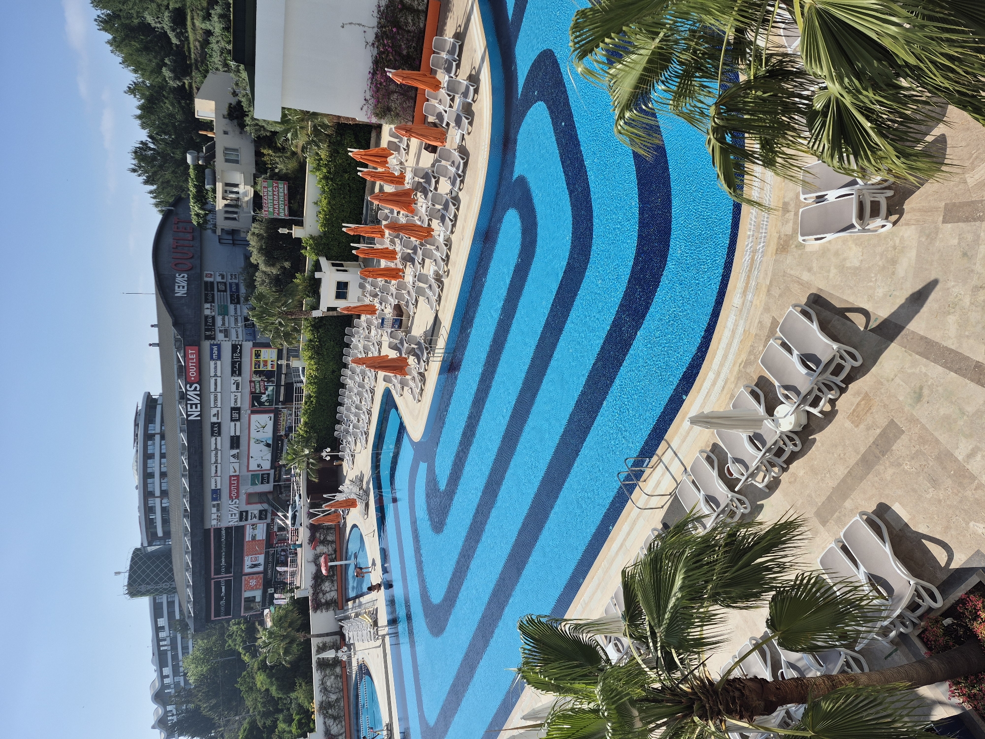 This is an aerial view of a resort swimming pool in Alanya, Turkey, identifiable by the "NEVİS OUTLET" shopping center and pharmacy in the background. The long, meandering pool is patterned with light and dark blue stripes. Rows of lounge chairs, mostly white with some orange shades, line the poolside. Palm trees are near the pool. A few people are visible in the water. The pool is surrounded by a tiled surface.