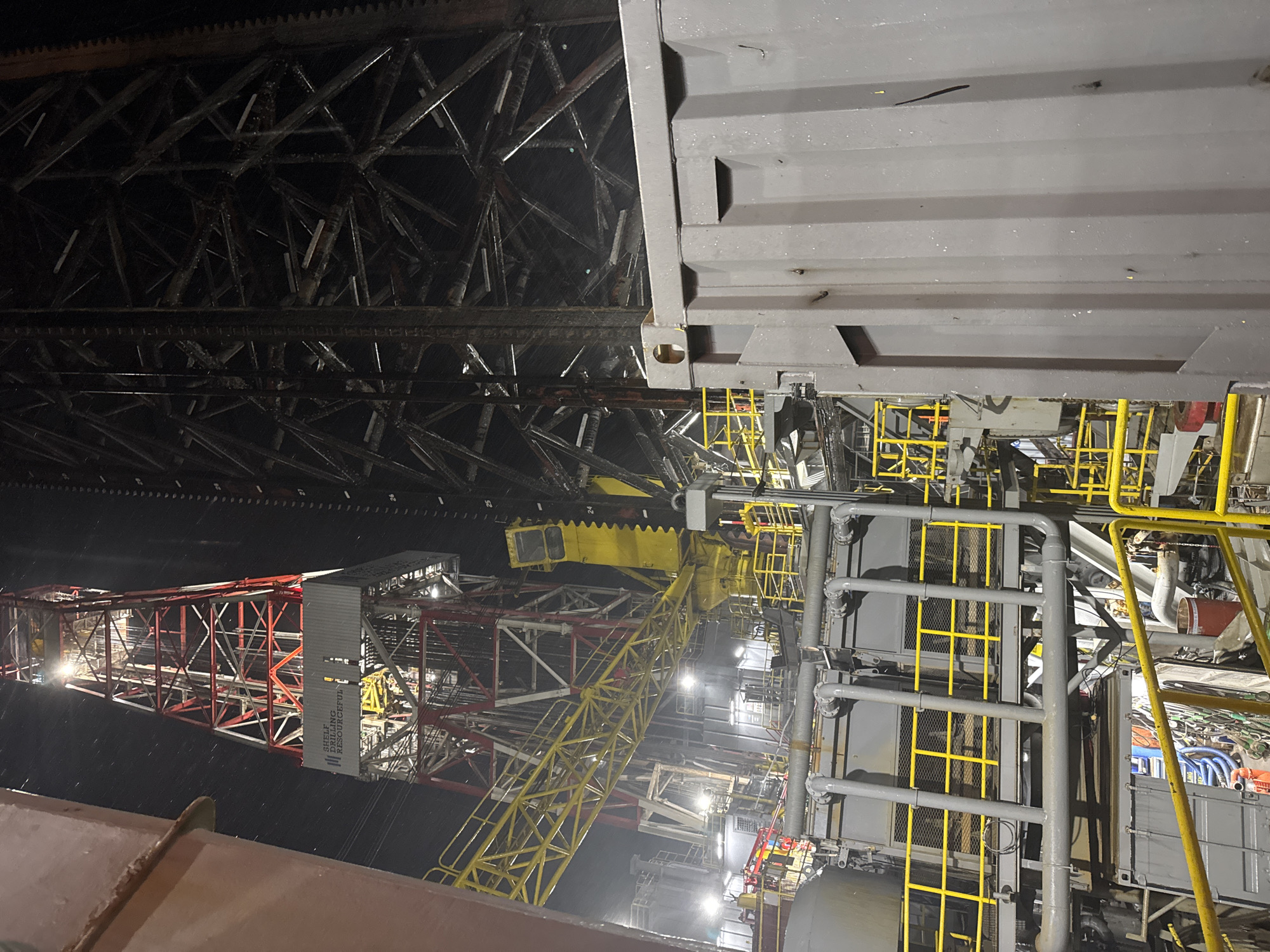 The image is a nighttime photograph of an offshore drilling rig. The rig's derrick, painted red and white, is prominently visible, illuminated by spotlights. Below the derrick, a complex network of metal walkways, pipes, and industrial equipment is arranged. The scene is industrial and appears to be a functional working environment, possibly at sea or in a port. The overall impression is one of a large-scale, operational piece of heavy machinery.