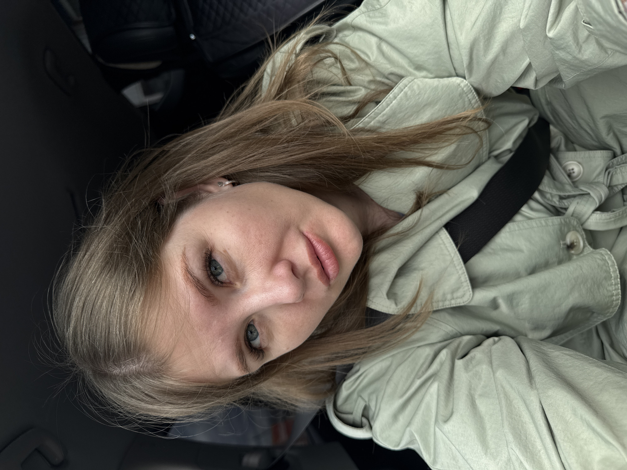 A diagonal portrait captures a young woman inside what appears to be a car. She has light brown hair, blue-green eyes, and fair skin, looking directly at the camera with a neutral expression. She is wearing a light green or sage-colored trench coat, with a black seatbelt positioned across her chest and shoulder. Visible details include a small heart-shaped earring in her left ear and a silver-colored ring on one of her fingers. The background is dark and out of focus, consistent with a vehicle's interior. The lighting suggests it is daytime. No specific activity beyond posing for the photo is evident, and no text is visible.