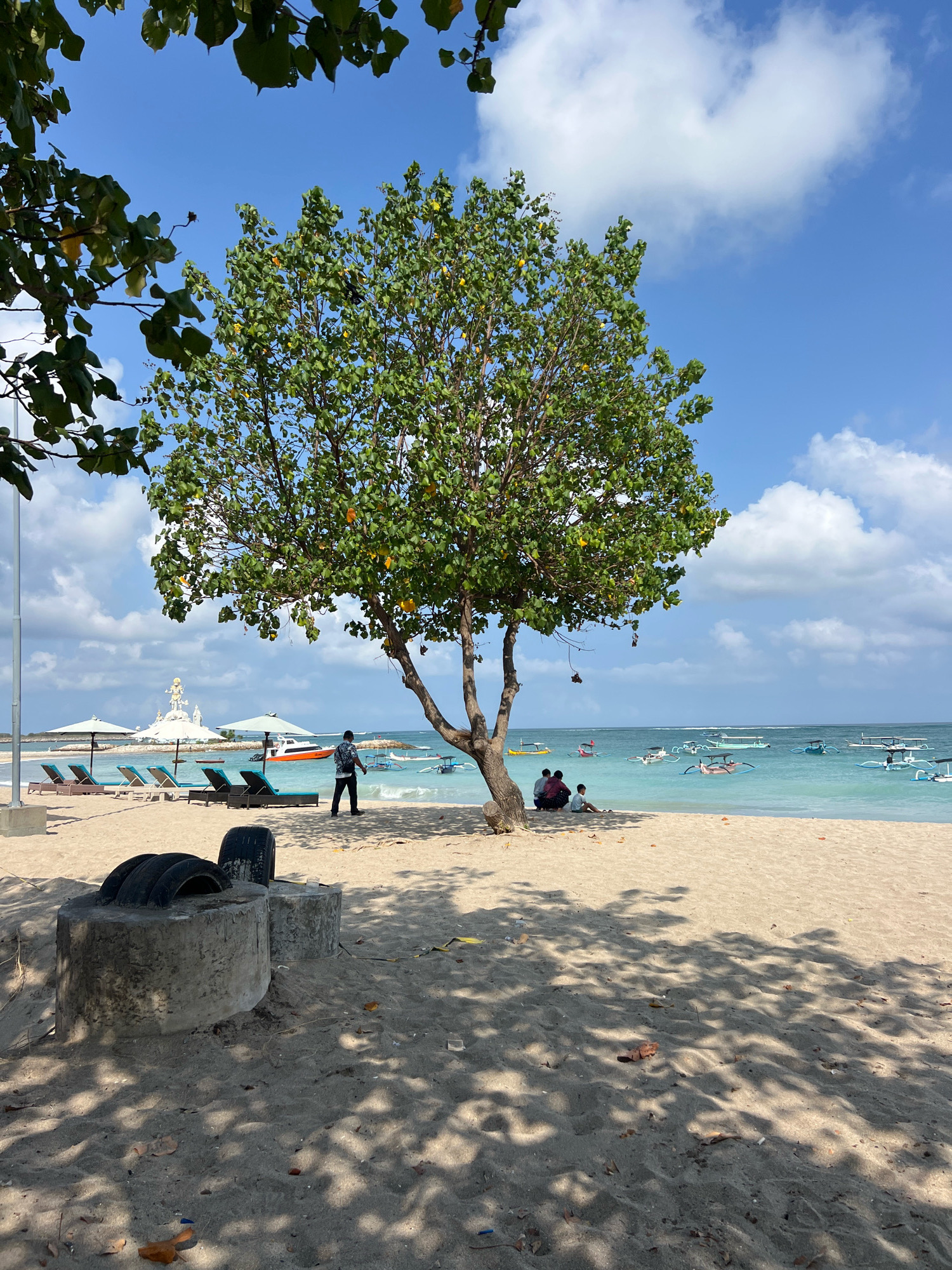 This image depicts a sunny day at Kuta Beach in Bali, Indonesia. A large tree with green leaves and some yellow fruit stands prominently in the foreground, casting dappled shadows on the sandy beach. To the left, several empty lounge chairs and umbrellas are arranged on the sand, indicating a resort or public beach area. In the middle ground, a person wearing a dark shirt and pants walks along the shoreline. Further out in the turquoise water, numerous small, traditional Balinese fishing boats with colorful outriggers are visible. In the distance, a large white statue, likely a religious monument, can be seen on the left side of the image. The sky is a clear blue with scattered white clouds. In the foreground, two concrete structures topped with old tires are partially visible on the sand. A small group of people are seated on the sand under the shade of the tree, to the right of the tree.