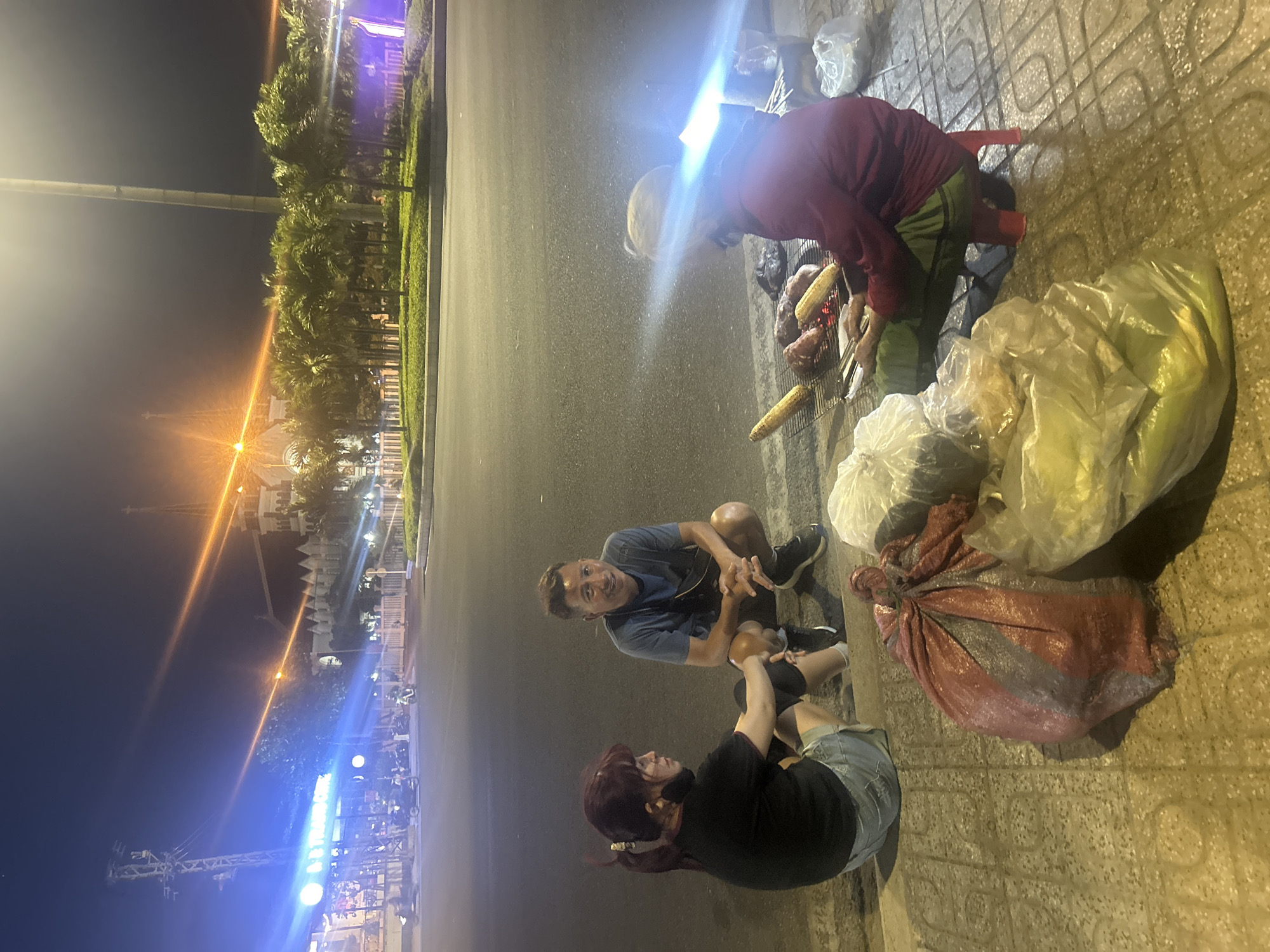 The image shows an outdoor scene at night in what appears to be a street in Bắc Nha Trang, Vietnam. A vendor with a headlamp is grilling corn and sweet potatoes on a small charcoal grill. Next to the vendor are several plastic bags containing cooked food, and a red and gray sack. A man and a woman are kneeling and talking to each other nearby. The street is paved, and there are palm trees and buildings with lights in the background. The sky is dark.