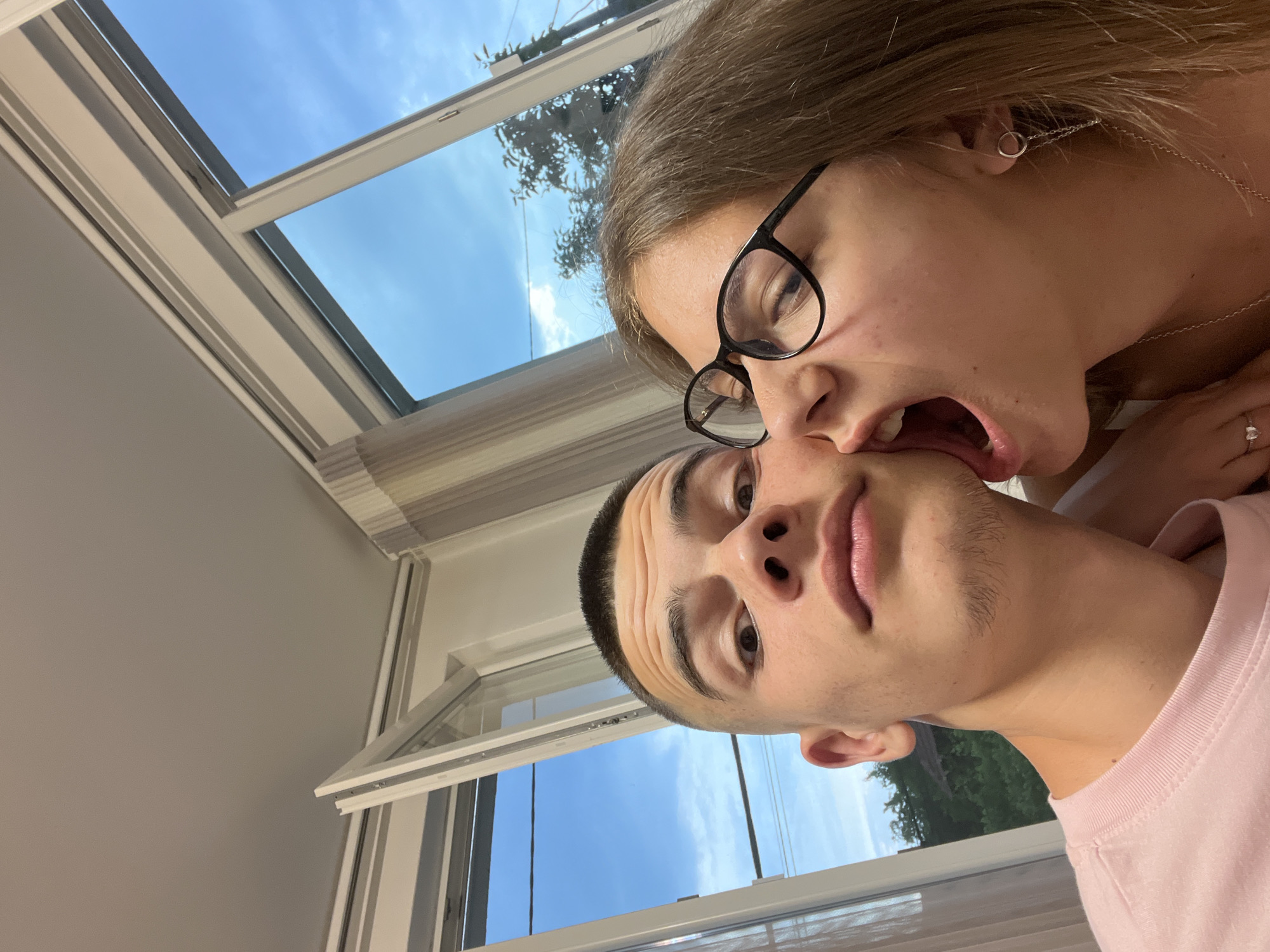 The image captures two young individuals in an indoor setting in Karavelovo, Bulgaria. A male, with a buzz cut and wearing a light pink crew-neck t-shirt, is positioned on the left, looking directly at the camera with wide eyes and visible forehead wrinkles. To his right, a female wearing black-framed glasses and brown hair has her mouth wide open, her tongue extended towards the male's right cheek/jawline in a playful interaction. She wears a silver hoop earring with a chain, a silver necklace, and a heart-shaped ring on her left hand, which rests on the male's shoulder. The background reveals white-framed windows, some partially open, showing a clear blue sky with scattered clouds, green trees, and utility wires, indicating an outdoor view. Sections of light-colored striped blinds or curtains are visible on the right and left, and a plain light ceiling occupies the top of the frame.
