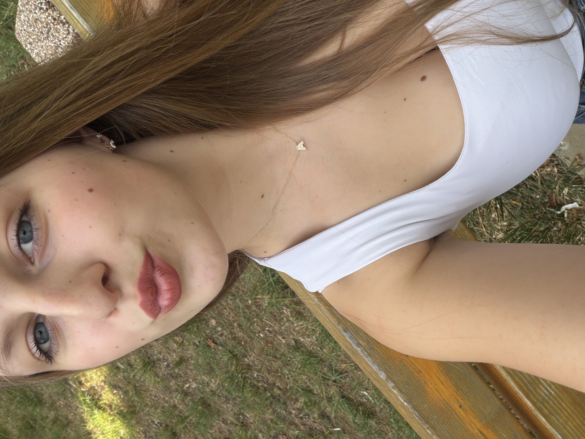 A young woman with long, brown hair and blue eyes is taking a selfie outdoors, sitting on a wooden bench. She is wearing a white top and a gold necklace with a small letter "A" pendant. Her lips are pursed. The background consists of green grass and some gravel.