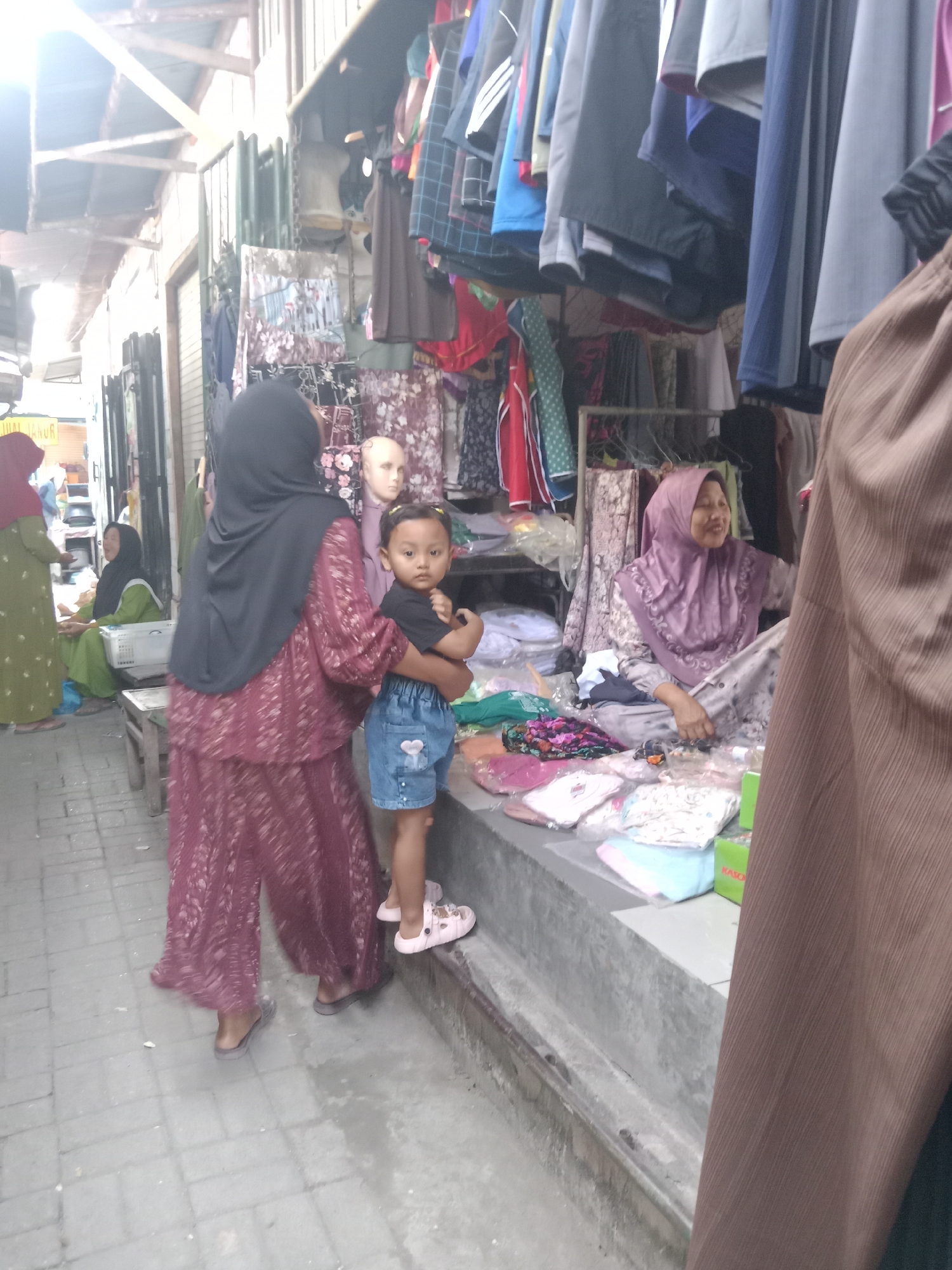 This image depicts an outdoor market scene in Klepek, Indonesia. In the foreground, a woman wearing a dark hijab and a maroon patterned dress stands holding a young child. The child is wearing a black t-shirt and blue denim shorts. To the right, another woman, also wearing a light purple hijab, sits behind a display of clothes and textiles. She is smiling and appears to be a vendor. Several clothing items are visible, including dresses, shirts, and patterned fabrics, displayed on mannequins and racks. The market area is paved with bricks, and there are wooden structures and awnings overhead, suggesting a covered market. In the background, other individuals and stalls are partially visible. The overall atmosphere appears to be a typical bustling marketplace.