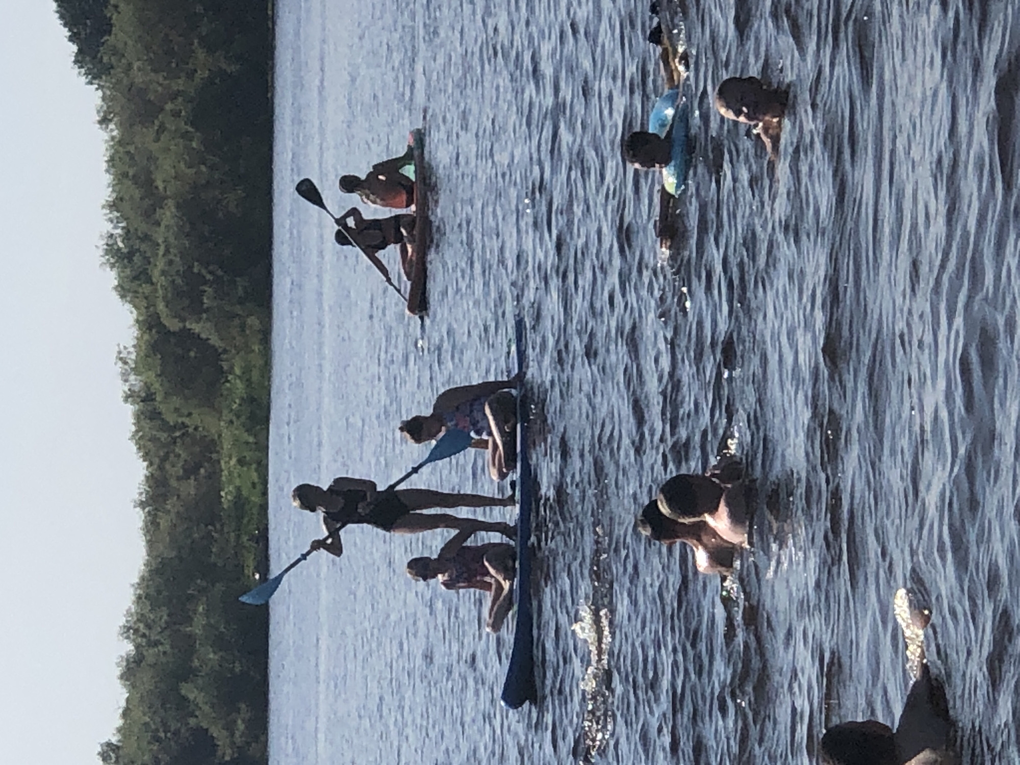 The image shows a body of water with several people engaged in various water activities under a clear sky. In the foreground, several individuals are swimming. One person is partially visible on the left, with their head and shoulders out of the water, holding a shiny object. Further back and to the right, two people are swimming, with one wearing a blue inner tube. Another person's head and shoulders are visible in the middle right, looking towards the camera.
In the midground, two people are on a paddleboard. One is standing and holding a paddle, while the other is seated. Behind them, another paddleboard carries two individuals, both seated.
The background consists of a dense line of trees and vegetation along the shoreline, with a faint, hazy horizon. The water is dark blue and appears to be moderately choppy, with ripples and small waves. The overall impression is a sunny day spent outdoors in a natural setting. There is no indication of the city of Solikamsk, Russia.