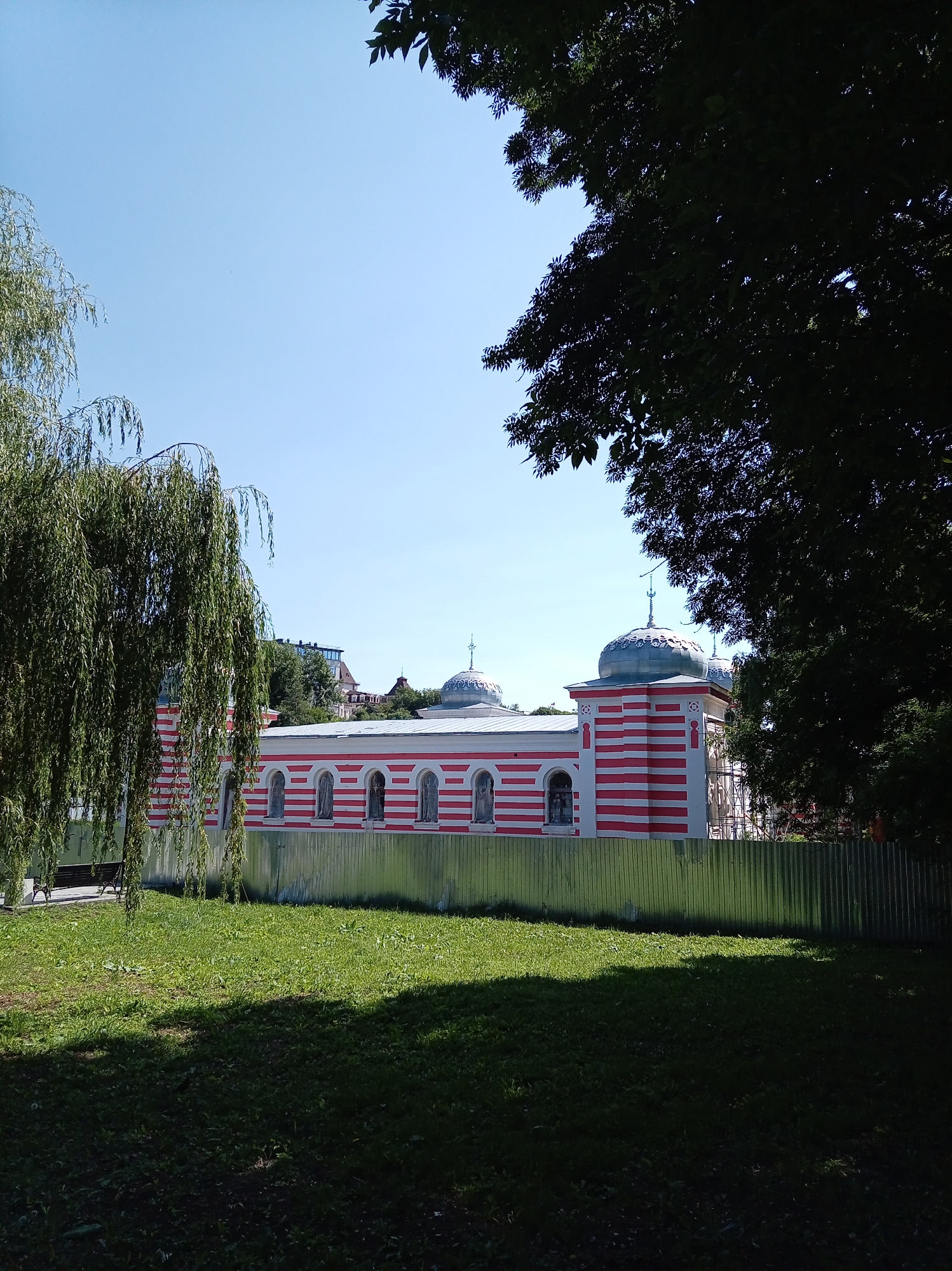 The image depicts a building with red and white striped walls and dome-shaped roofs. The building is fenced off with a corrugated metal fence. In the foreground, there is a grassy lawn, and in the background, there are trees and other buildings. The sky is clear and blue. The building appears to be the "Grotto of the Mermaid" located in the city of Zheleznovodsk, Russia.