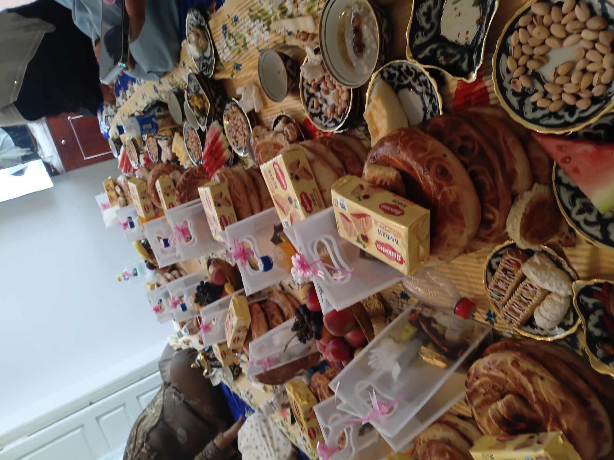 The image depicts a table laden with food and drinks, likely for a gathering or celebration. There are various types of bread, including large, circular loaves with intricate patterns, and smaller, crescent-shaped pastries. Several plates contain nuts, some appearing to be pistachios. There are also bowls of fruit, including grapes and peaches, and slices of watermelon. Packaged snacks, possibly cookies or biscuits, are stacked on the table, some with prominent branding. Bottles of liquid, possibly juice or water, are also present. Decorative elements like small golden teapots and translucent gift bags with pink ribbons are visible. The table is covered with a patterned cloth. The background suggests an indoor setting, with hints of furniture and a wall. The scene appears to be from a city in Kyrgyzstan, specifically Osh. The overall impression is one of abundance and hospitality.