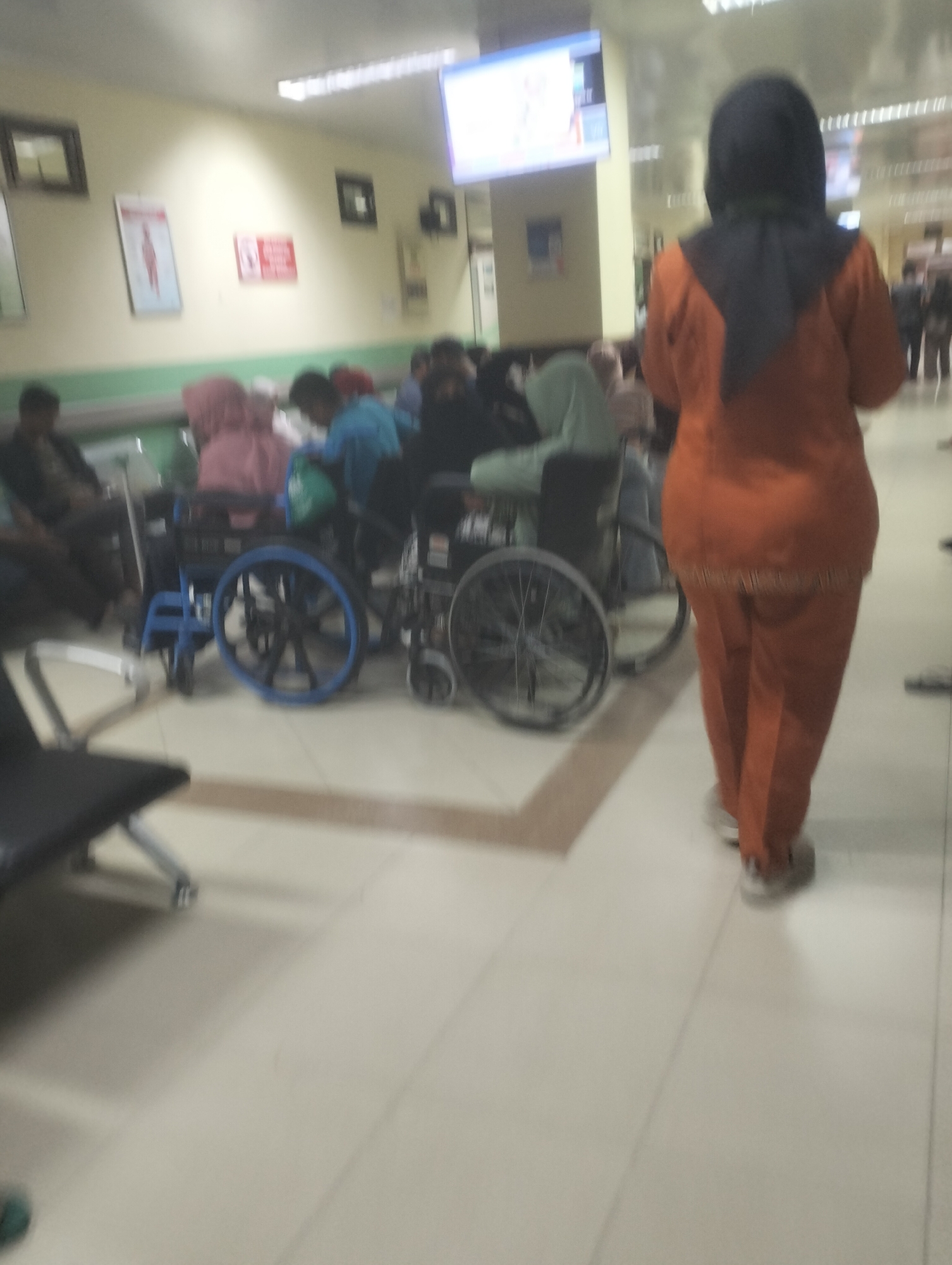In an indoor waiting area, likely a hospital or clinic, located in Air Jamban, Indonesia, several individuals are present. A person wearing an orange uniform and a dark hijab is walking away from the camera. In the background, multiple people are seated in wheelchairs and on benches. One wheelchair is prominently displayed with blue accents on its wheels. The environment appears to be a well-lit, tiled corridor. There are posters on the wall, and a large screen is visible on the ceiling. The overall scene depicts a common waiting area, possibly for medical attention.