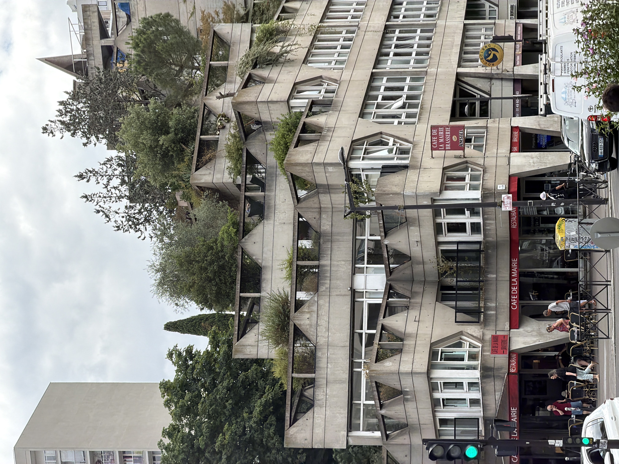 This image captures a street-level view of the "Le Grand ensemble des Bords de Seine" apartment complex in Ivry-sur-Seine, France. The brutalist architecture of the building dominates the frame, characterized by its exposed concrete, geometric protrusions, and extensive use of balconies and terraces integrated with greenery. Several businesses are visible at the ground level, including "Café de la Mairie Brasserie" and a "Restaurant," with outdoor seating areas set up on the sidewalk. People are present, some seated at the cafe tables, others walking by or standing near the entrance. A traffic light with a green signal is visible on the left, along with street signs and a parked black Audi sedan. In the background, a tall, plain residential building stands to the left, and trees and vegetation are interspersed throughout the complex, softening the harsh concrete. The sky is overcast, suggesting a cloudy day.
