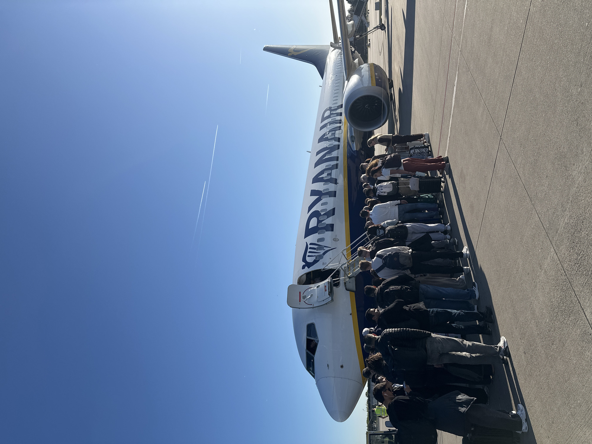 This sunlit image captures a long queue of passengers boarding a Ryanair aircraft on an airport tarmac in Weeze, Germany. The perspective is taken from slightly behind and to the side of the queue, looking towards the front of the airplane.
The main subjects are a large group of people and a white and blue Ryanair airplane, likely a Boeing 737, with "RYANAIR" clearly visible along its fuselage and the "RYR" logo on the tail. The passengers, dressed in casual attire and carrying various bags and luggage, are lined up in single file, extending from the front door of the aircraft where a set of mobile stairs is connected. They appear to be waiting to ascend the stairs and enter the plane.
The scene is an airport apron under a bright, clear blue sky. Strong sunlight casts prominent shadows of both the aircraft and the people onto the asphalt tarmac, indicating a sunny day, likely during midday or early afternoon. Several thin, white contrails are visible high in the sky. The ground shows typical airport markings. No strong emotions are discernible from the passengers, who seem to be engaged in the routine process of boarding.