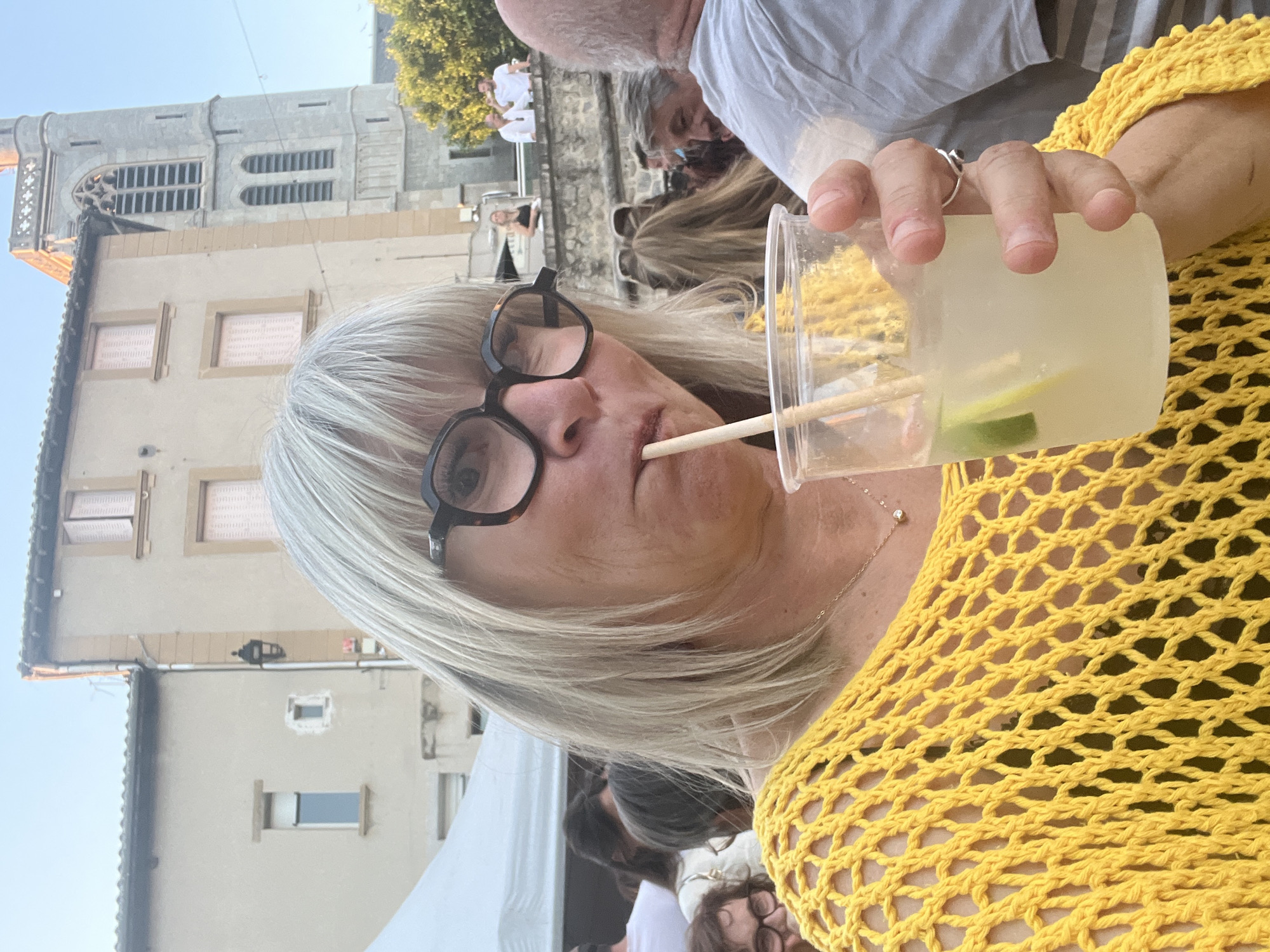 In Saint-Galmier, France, a woman with short, blonde hair and glasses is captured in a selfie as she drinks from a clear plastic cup containing a pale yellow liquid with lime slices and a wooden straw. She is wearing a vibrant yellow, crocheted top that reveals a black garment underneath, and a delicate necklace. The background is softly blurred, showing a street scene with buildings, including a stone tower that resembles a church steeple, and a white tent structure. Other individuals are visible in the background, some blurred, suggesting an outdoor gathering or event. The overall lighting indicates it is daytime, likely late afternoon, with the sun casting warm light on the scene.
