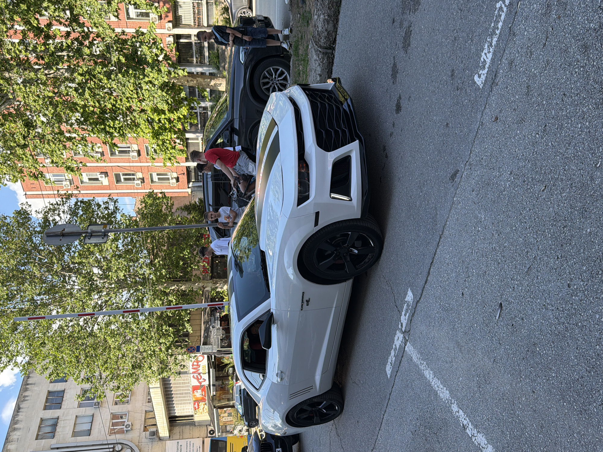 The image shows a white Camaro 6.2L parked in a parking space in front of the "Tempo" pizzeria in Pleven, Bulgaria. Several people are gathered near the car and the SUV parked next to it. The background features a brick building and a tree. A parking barrier is also visible in the frame.
