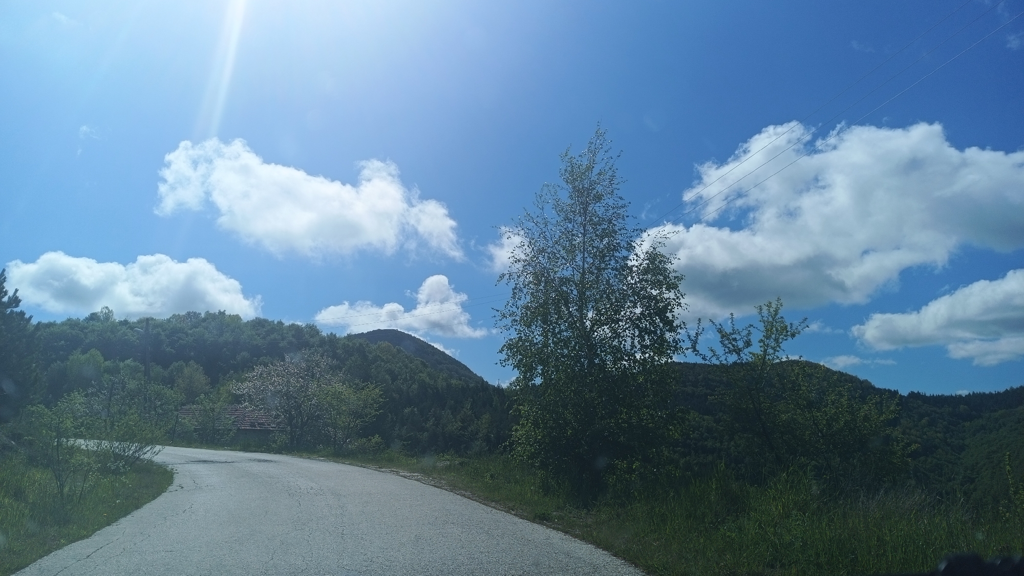 The image shows a winding paved road in a rural area, likely near or in the vicinity of Vrata, Bulgaria based on request. The road curves to the left in the foreground. Lush green vegetation covers the slopes of hills in the background, which are partially obscured by a tall, slender tree located to the right of the road and another to the left. In the distance, a mountain peak is visible. Scattered clouds are present in the bright blue sky, with the sun shining brightly, creating a lens flare effect. Power lines can be seen stretching across the sky in the mid-right portion of the image. A building partially hidden by trees can be seen on the left side of the road further down the road.