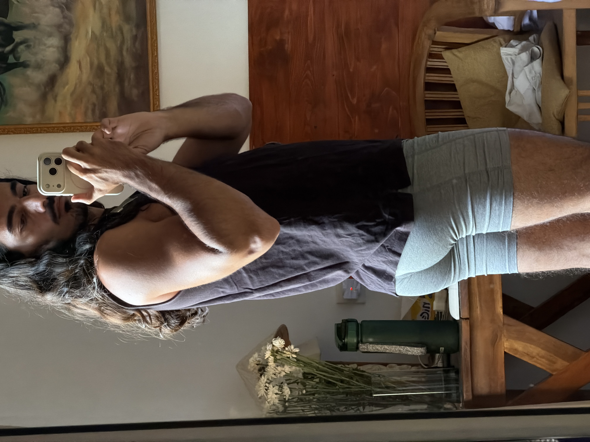 A man with dark hair and a beard is taking a selfie in a mirror. He is wearing a black tank top and grey shorts. In the background, there is a painting on the wall, a wooden cabinet, and a chair with pillows. There is also a vase of white flowers and a green object on a table in front of the mirror. The reflection suggests the location is in North Kuta, Indonesia, given the style of furniture and decor.