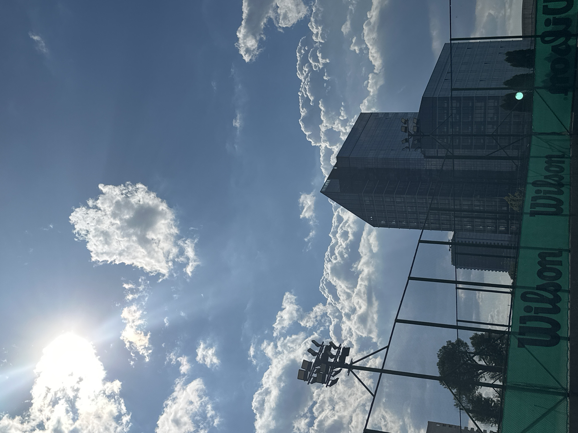The image shows a view of the sky with cumulus clouds and a building in Sofia, Bulgaria. The sun is visible through the clouds in the lower-left of the frame. The building is modern and appears to be a skyscraper. A tennis court with a green Wilson branded windscreen and a lighting rig is visible in the foreground. The perspective is skewed and appears to be from a low angle.