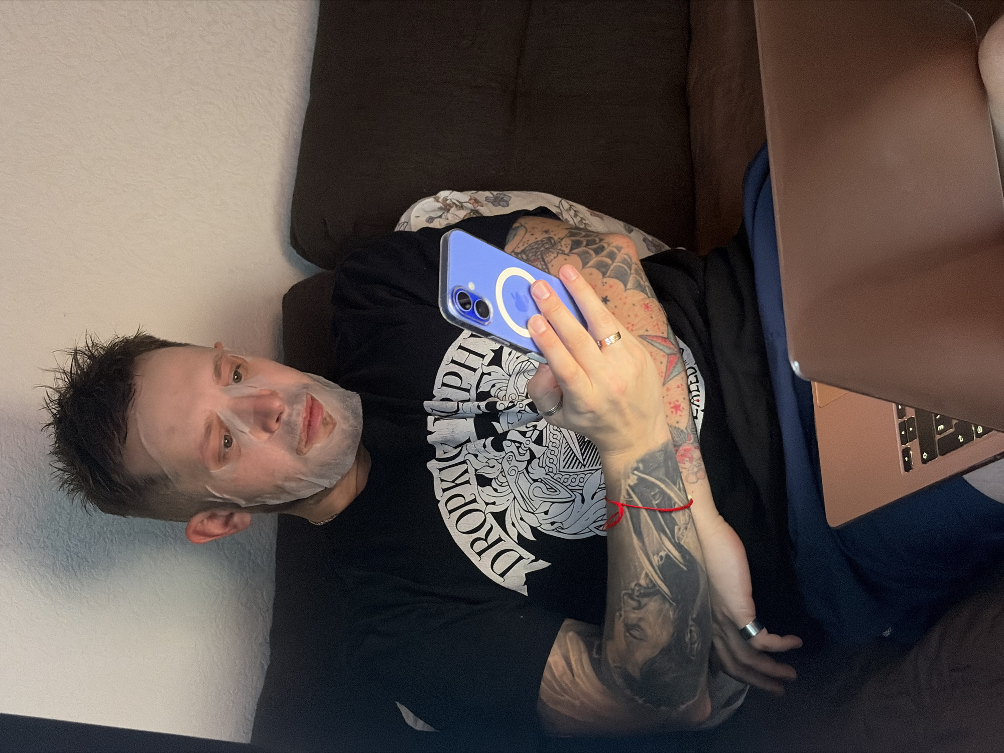A person is lying on a dark brown couch, wearing a black t-shirt and a white face mask. They are holding a blue smartphone in their right hand and a rose-gold colored laptop is resting on their lap. The person has tattoos on their arms, and is wearing a ring on their left ring finger. The location cannot be determined from the given information.
