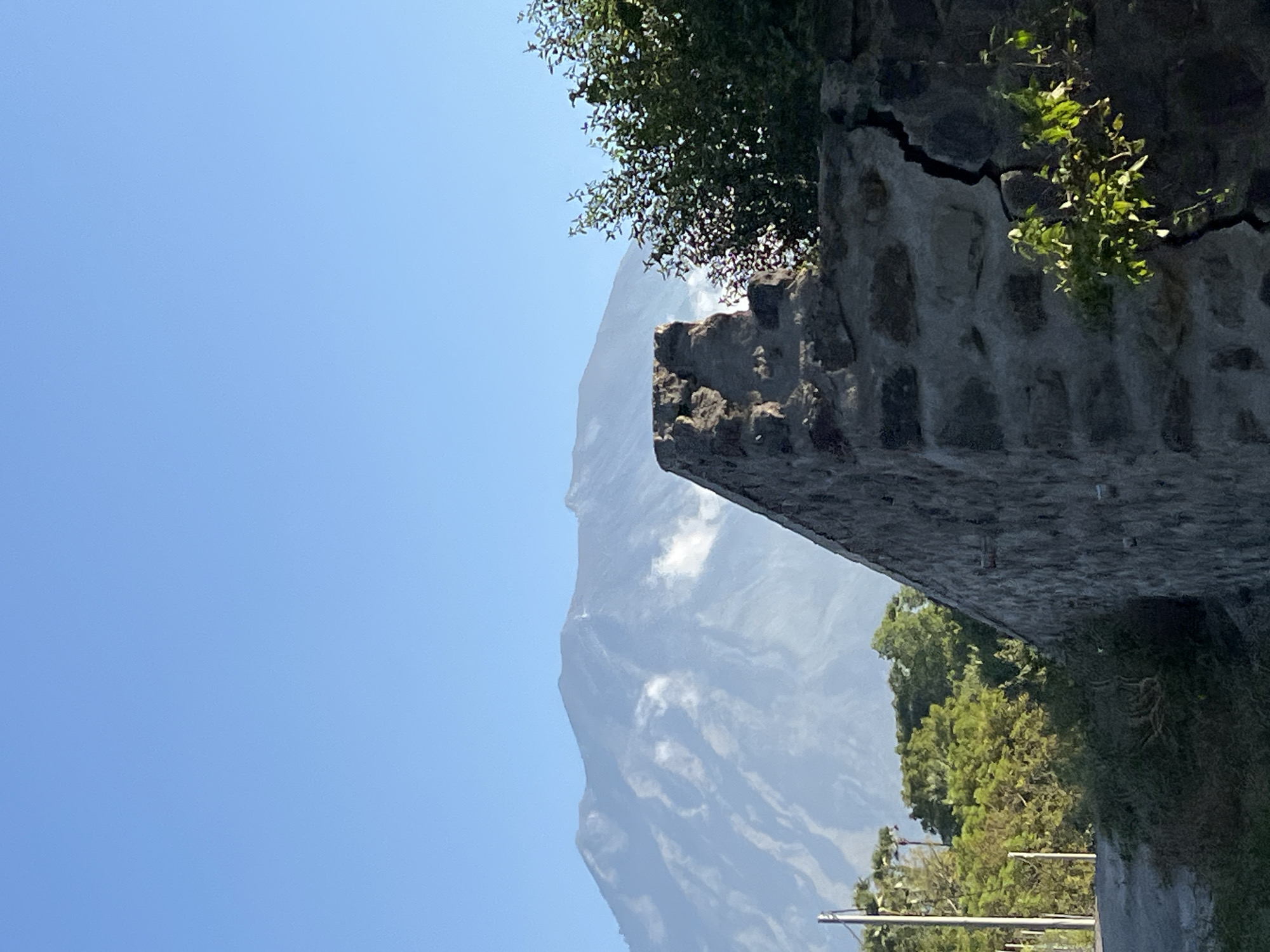 This media file is an image of a mountainous landscape in Kubu, Indonesia. The image is oriented vertically. In the foreground, there is a stone wall with a large crack running through it. Green foliage grows from the crack and from the top of the wall. Beyond the wall, a large, imposing mountain looms, its slopes partially obscured by soft white clouds. The sky is a clear, bright blue, suggesting a sunny day. In the lower right of the image, the tops of trees and what appear to be utility poles are visible, hinting at a road or pathway near the foreground. The scene conveys a sense of natural grandeur and perhaps the ruggedness of the terrain. There is no visible text in the image.