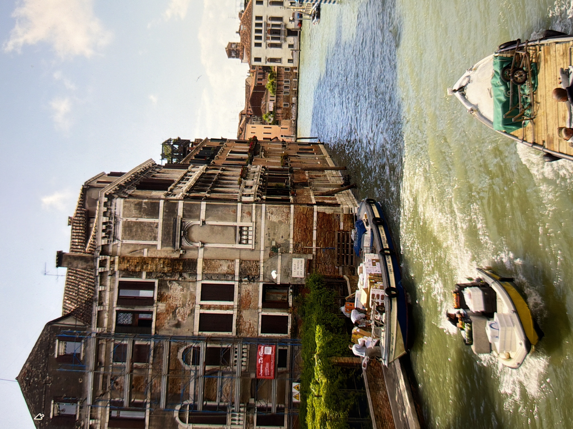 The image depicts a canal in Venice, Italy, not Kyiv, Ukraine. In the foreground, a small yellow boat with a white cabin navigates through the water, carrying crates and a person at the helm. Behind it, a larger, flat-bottomed boat is laden with numerous boxes and is manned by several individuals. This boat is docked alongside a canal, where a verdant green hedge lines the embankment. The backdrop is dominated by multi-story buildings with weathered facades, showcasing a mix of architectural styles. Some buildings are under renovation, indicated by scaffolding. The sky is partly cloudy and a bird is visible in flight.
