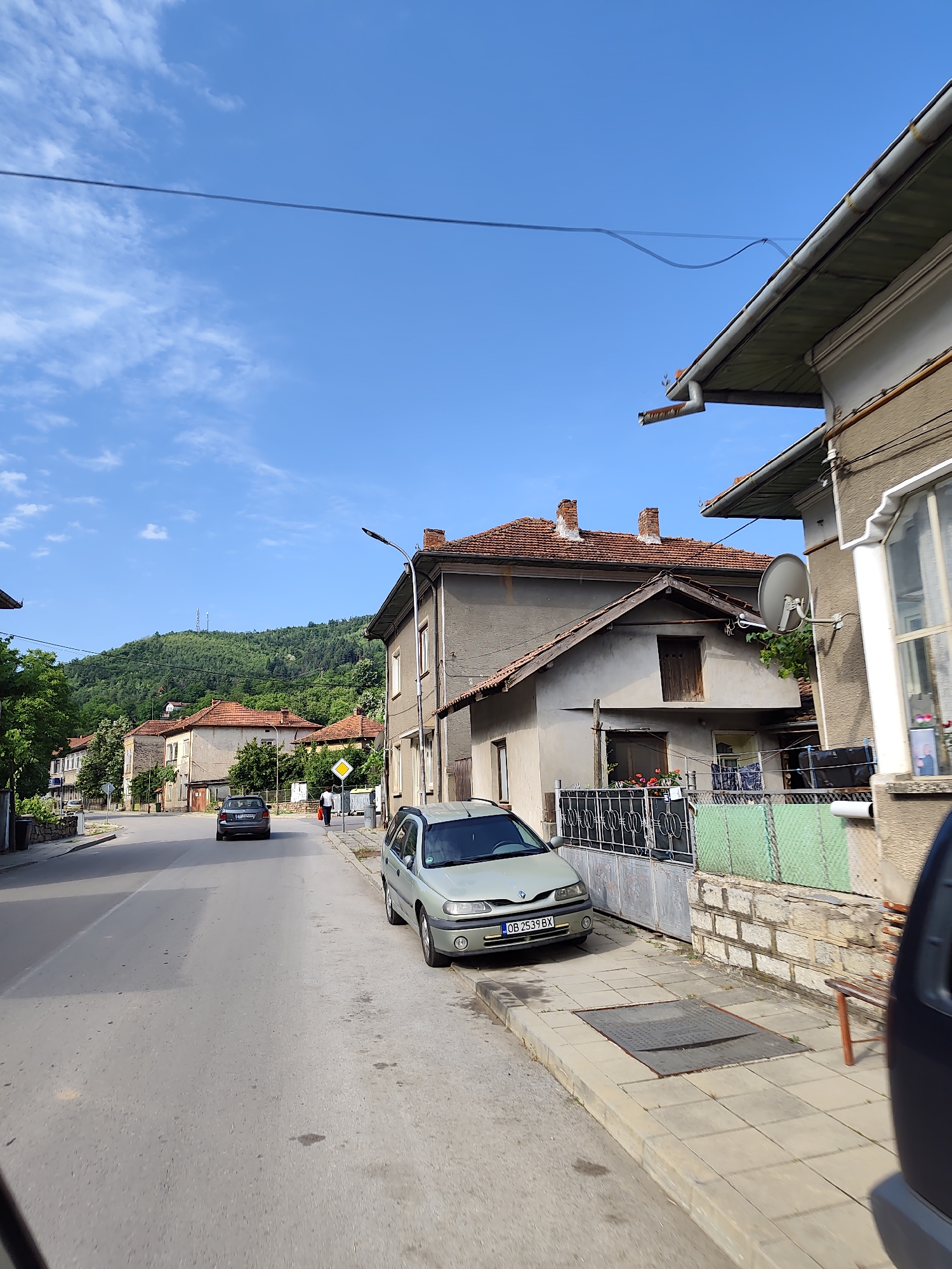 A street scene in the Bulgarian village of Lesidren shows a clear blue sky with scattered clouds and a gently sloping, tree-covered hill in the background. Two cars are visible on the paved road. A dark grey car is further down the street, and a lighter colored car is parked on the right side of the road. A person in white shirt and dark pants is walking away from the viewer on the sidewalk. Buildings with red tile roofs line the street, and a street lamp is positioned on the right side of the road. A satellite dish is mounted on the side of a building.