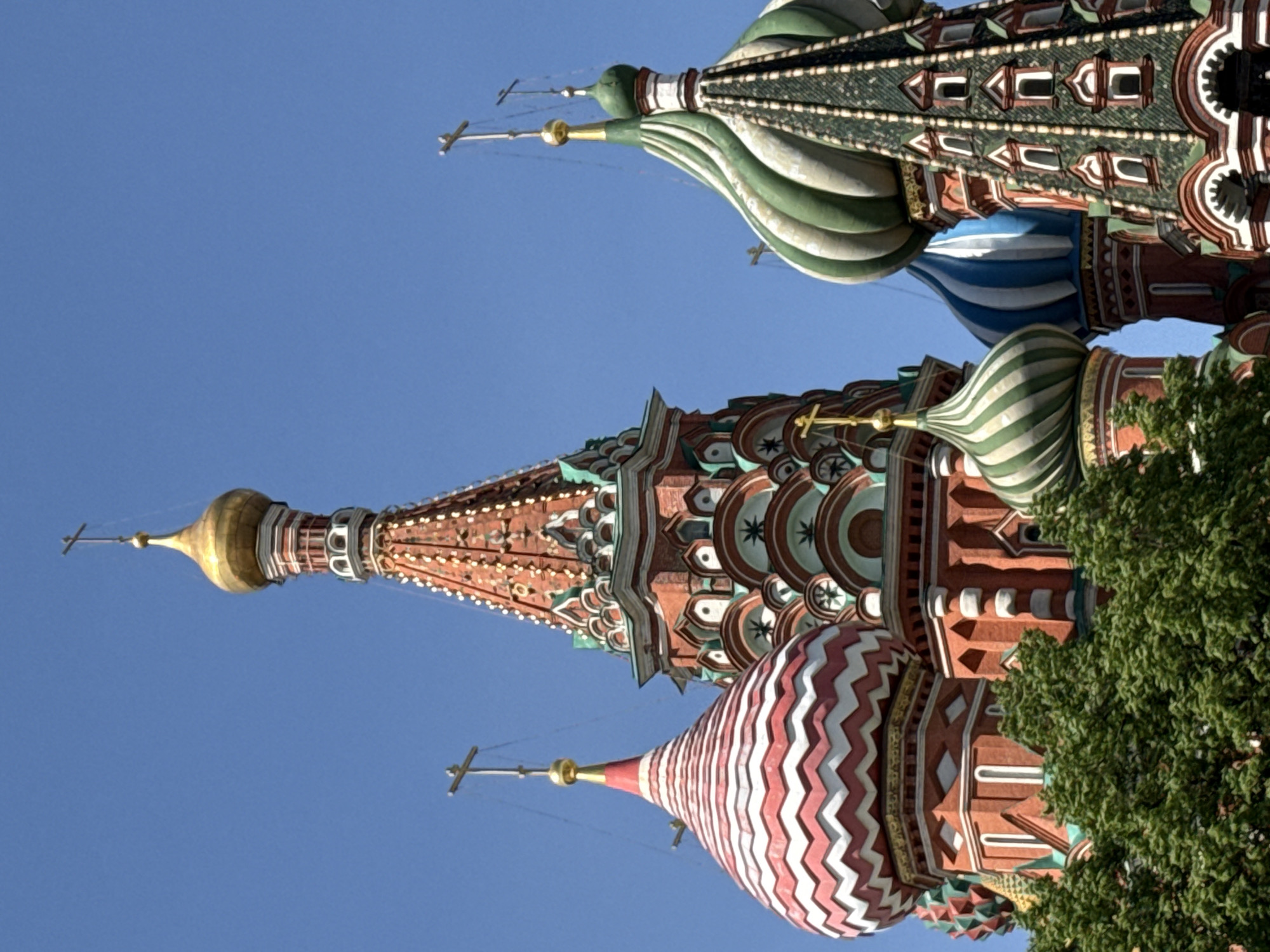 The image features a section of St. Basil's Cathedral in Moscow, Russia, viewed from a low angle. The sky is clear and blue. Several of the cathedral's distinct domes are visible, each with unique and elaborate designs. The closest dome has a red and white chevron pattern. The next dome has a multicolored pattern, dominated by red and green. The top-most dome visible features green and white stripes. All domes have golden crosses atop them. The lower right corner of the image is partially obscured by a green leafy tree. The image is rotated counter-clockwise.
