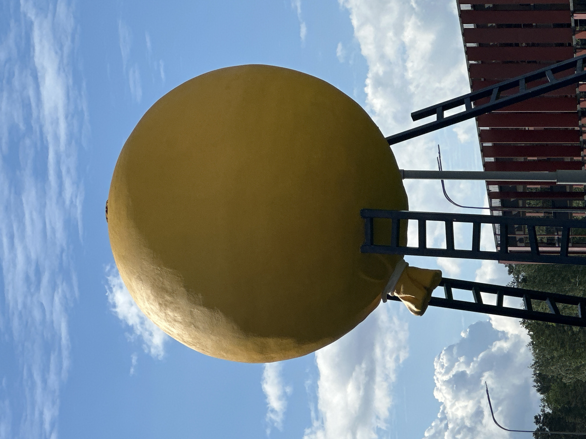 The image shows a large yellow spherical object, resembling a giant lemon, set against a blue sky with scattered white clouds. The sphere appears to be part of a structure, as black ladders and a red and brown multi-layered building component are visible to the right. The presence of trees in the background further suggests the location is outdoors. The image is rotated, with the sky at the bottom. Given the location information provided (Бараўлянскі сельскі Савет, Belarus), it is likely a public art installation or a decorative element within the town.