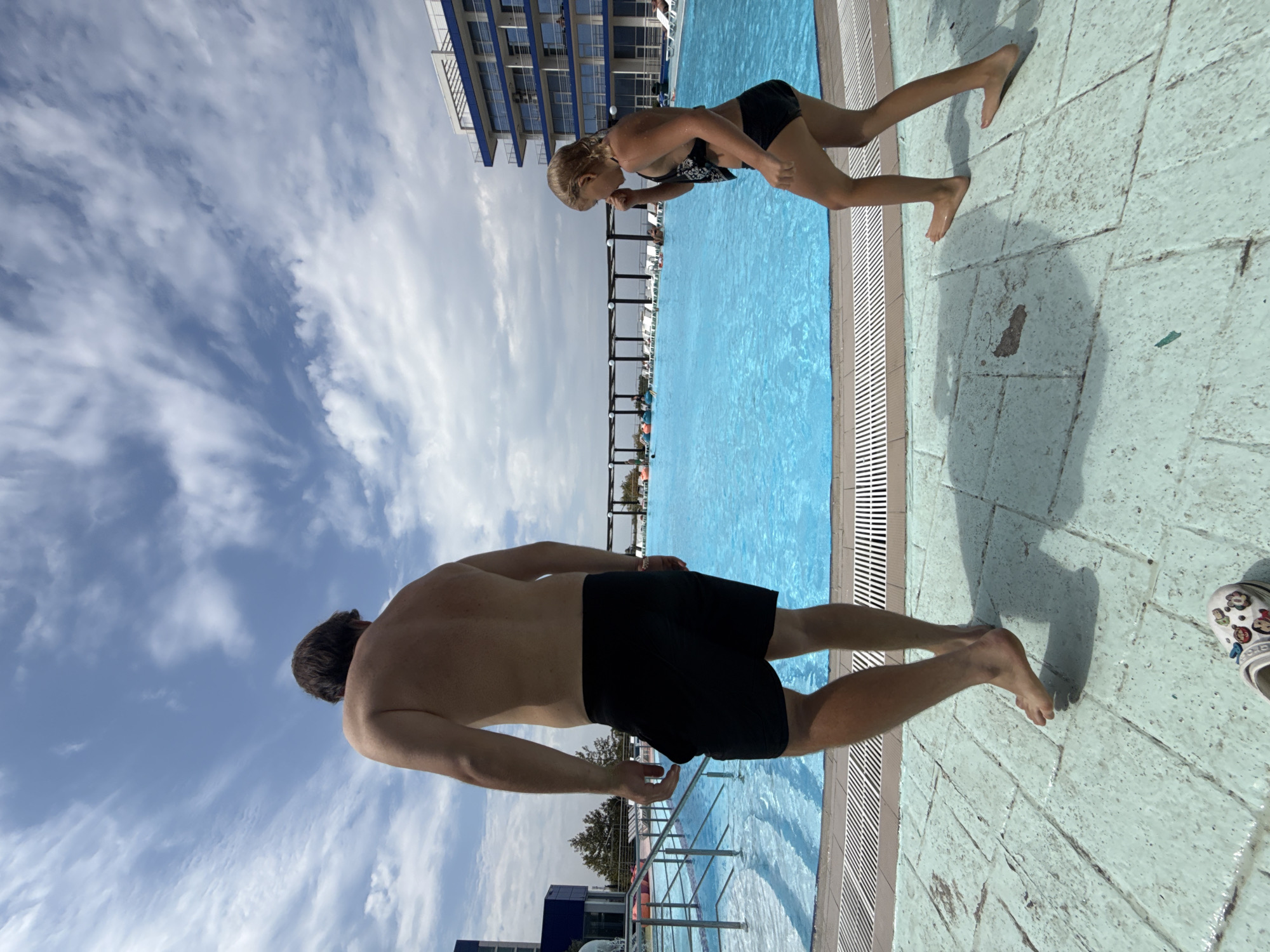 This is a horizontal, eye-level, wide shot taken on a sunny day, likely in the afternoon, at an outdoor swimming pool. The sky is a bright blue with scattered white, wispy clouds. The pool water is a vibrant blue and appears still.
In the foreground, a man is positioned with his back to the camera, looking towards the pool. He has short brown hair and is wearing black swim trunks. His body is muscular and his skin is tanned.
In the mid-ground, a young girl with blonde hair, wearing a black bikini, is standing at the edge of the pool. She is leaning forward, with her hands on her knees, and appears to be looking down at something. Her stance suggests she might be preparing to jump or is in the process of some movement.
The setting includes a concrete poolside with visible tile patterns. To the left of the frame, a modern, multi-story building with blue and white accents is visible, suggesting a hotel or resort. Further in the background, there are structures that look like covered seating areas or pergolas, common at swimming pools.
The overall mood seems relaxed and summery, consistent with a day at a swimming pool. There is no visible text in the image. The presence of both a man and a child implies a family or recreational outing. The lighting indicates it is daytime with clear skies, ideal for swimming.
