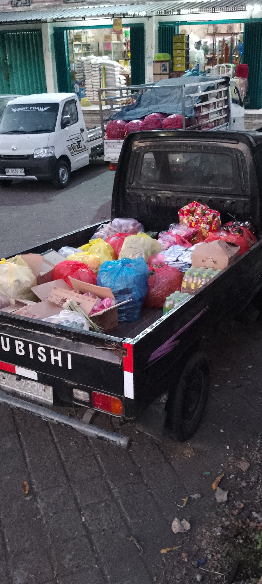 In Pekutatan, Indonesia, a black Mitsubishi pickup truck is parked on a paved surface, likely a street or parking lot. The truck bed is filled with various items, predominantly large, colorful plastic bags in red, yellow, and blue, suggesting they contain goods or waste. Several cardboard boxes are also visible, some open, revealing contents that appear to be packaged items and possibly produce. A small white Isuzu truck is parked in front of the Mitsubishi, and in the background, a building with green accordion doors is visible, with stacks of white bags resembling rice or other goods inside. The overall scene depicts a common urban or market setting, potentially a distribution or collection point.
