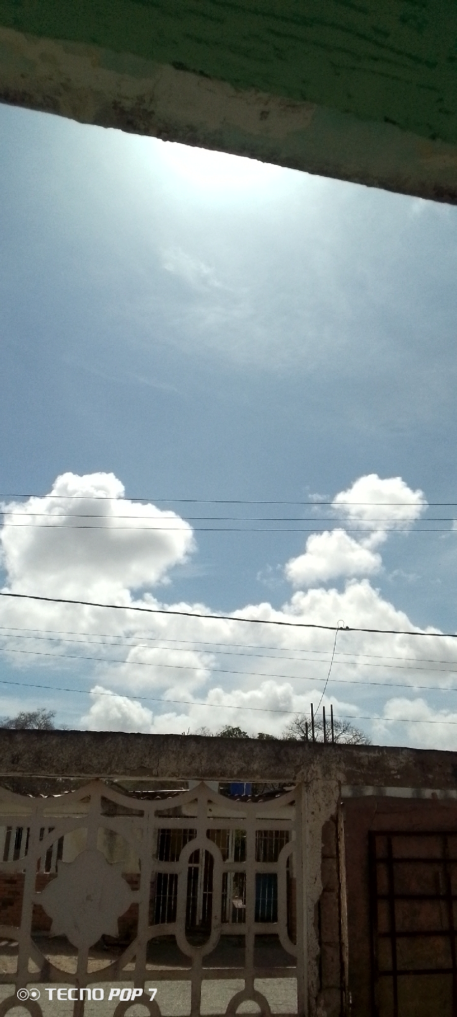The image, captured by a TECNO POP 7, presents a partially obscured view of a bright, daytime sky in Altagracia, Venezuela. The sky is predominantly blue, dotted with several white cumulus clouds. High overhead, the sun shines brightly. Electrical wires bisect the view horizontally at two points. The foreground is dominated by a concrete perimeter wall topped with sparse vegetation, and a wrought-iron gate. The structures and vegetation suggest a residential or commercial area.