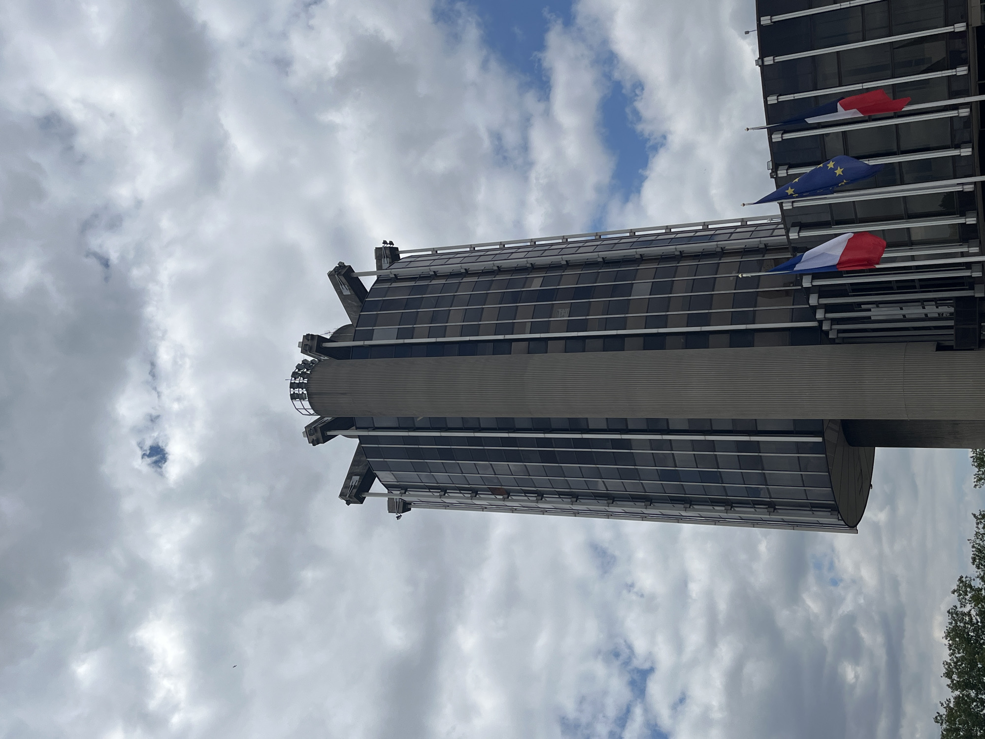 A low-angle shot captures a modern building against a cloudy sky. The building's facade is predominantly glass, reflecting the sky and clouds. Three flags are visible, mounted on poles extending from the building. Two of the flags appear to be the French tricolor, with blue, white, and red stripes. The third flag is the European Union flag, featuring a circle of yellow stars on a blue background.
The scene is set outdoors, with trees visible at the right edge of the frame. The weather is overcast, with a mix of gray clouds and patches of blue sky. The time of day is indeterminate due to the lighting conditions. The image appears to be taken in Créteil, France, as suggested by the provided location context. There are no people visible in the image, and the focus is on the architecture and the flags. The flags are slightly blurred due to movement in the wind, creating a dynamic element in the otherwise static scene. The building's structure is imposing, with a cylindrical section that narrows as it ascends.