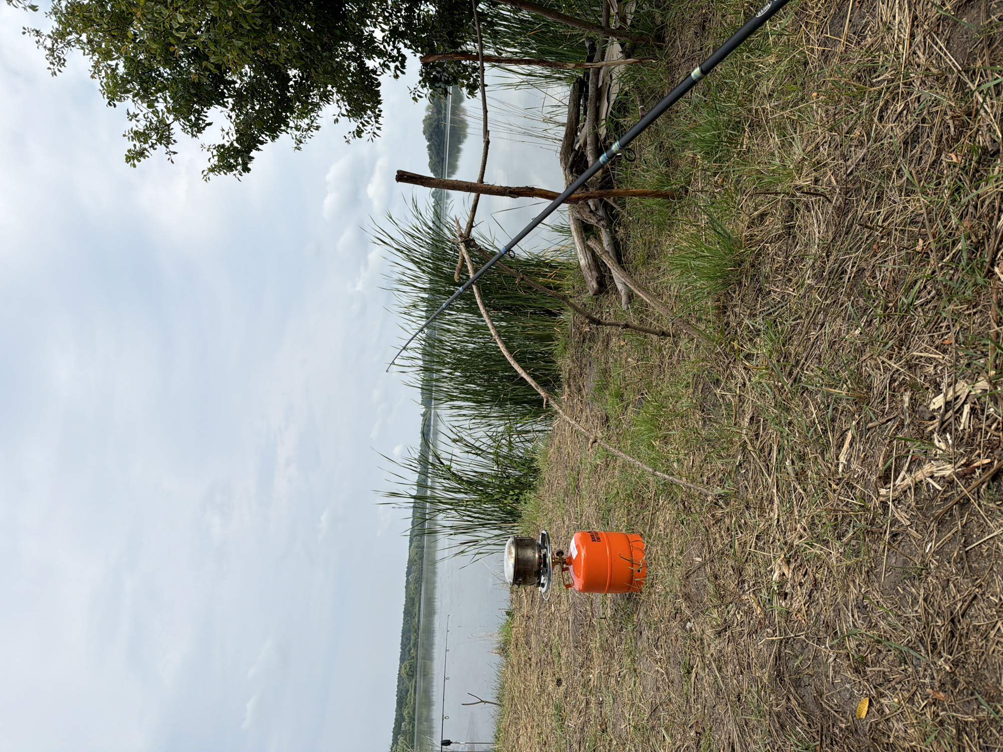 A gas camping stove with a pot on top sits on the dry grass near the water's edge in Ruda, Ukraine. The stove is orange and cylindrical, with a metal rack holding the pot. Behind the stove, tall reeds grow along the bank of a calm body of water, which is bordered by a distant tree line. A fishing rod is propped up on a wooden structure made of branches. The sky is overcast.