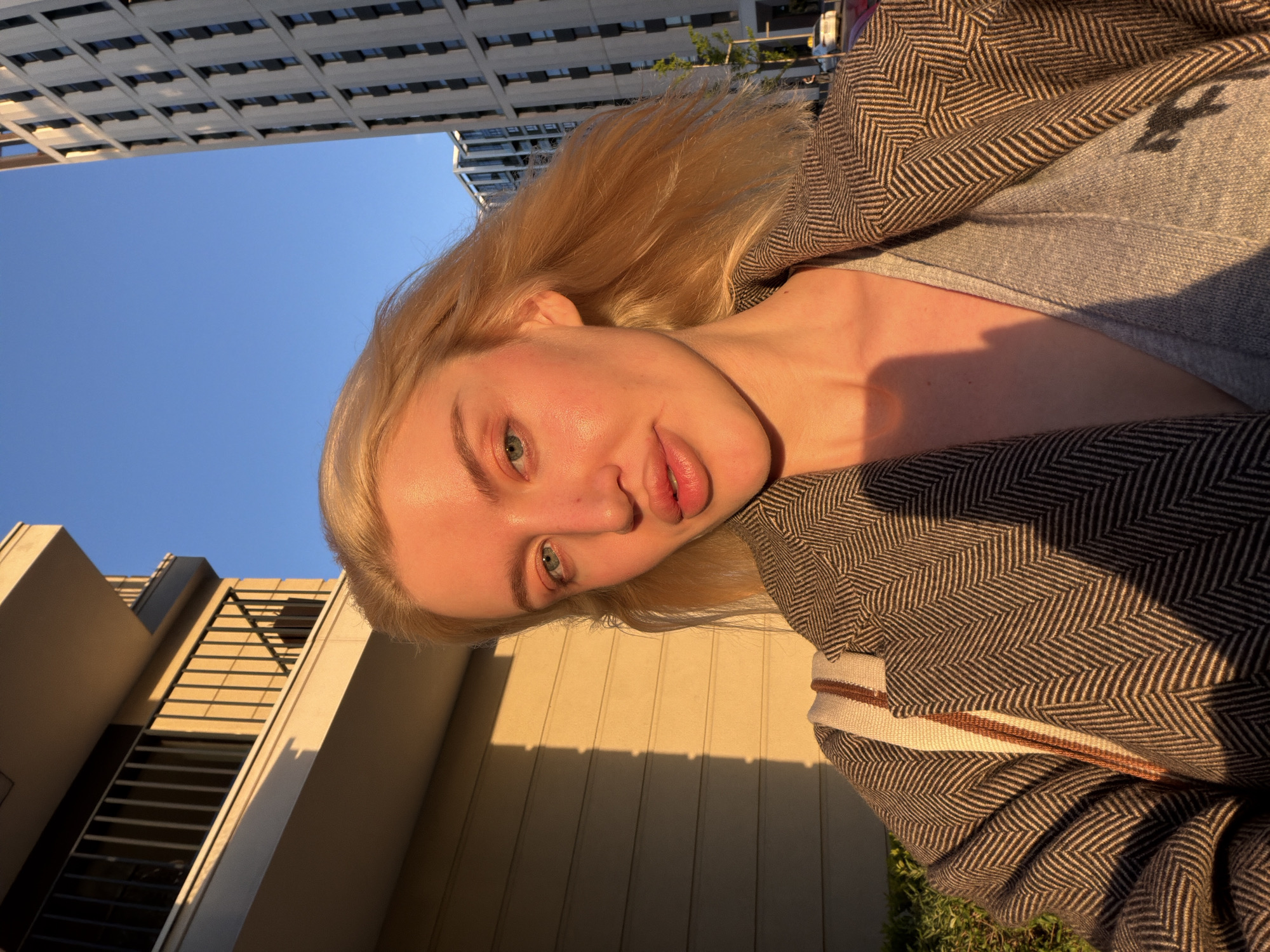 This image is a close-up, rotated portrait of a young woman taken outdoors in an urban setting. The woman has fair skin, blonde hair, and light-colored eyes. Her face is directly illuminated by warm, strong sunlight, highlighting her features and casting a bright glow on one side of her face, with soft shadows on the other. She looks straight into the camera with a neutral to slightly smiling expression, her lips slightly parted.
She is wearing a grey top, partially visible, beneath an open jacket or coat featuring a prominent brown and off-white herringbone pattern. A striped strap, likely from a bag, is visible over her shoulder.
The background shows a clear blue sky above and behind her. In the upper portion of the frame, there are modern, tall buildings with grid-like facades, appearing structurally upright despite the image's rotation. The lower background features a building wall or facade with horizontal elements, where long, distinct shadows are cast by architectural features and potentially foliage, indicative of direct sunlight. A hint of green foliage is visible at the bottom.
The strong, directional sunlight and the warm tones suggest the photograph was taken during golden hour, either in the late afternoon or early morning, on a sunny day. The setting is an outdoor urban environment, consistent with Belgrade, Serbia. No visible text is present in the image.