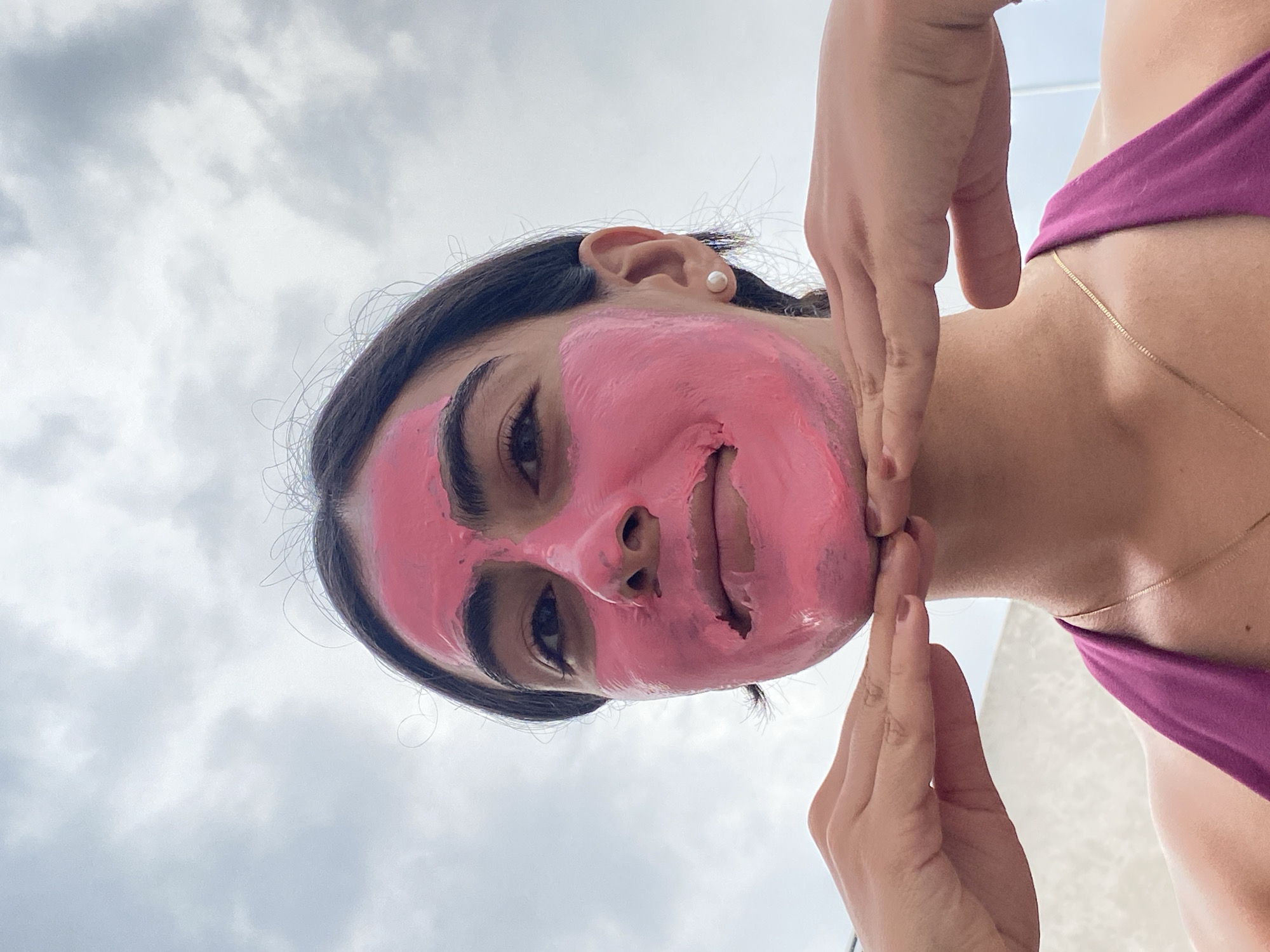 A woman wearing a pink face mask is holding her cheeks with her hands in an upward angle shot. She is wearing a magenta top and a gold necklace. The background shows a cloudy sky. The location appears to be outdoors, but it's not possible to determine if it is in Puebla, Mexico, based on visual information alone.