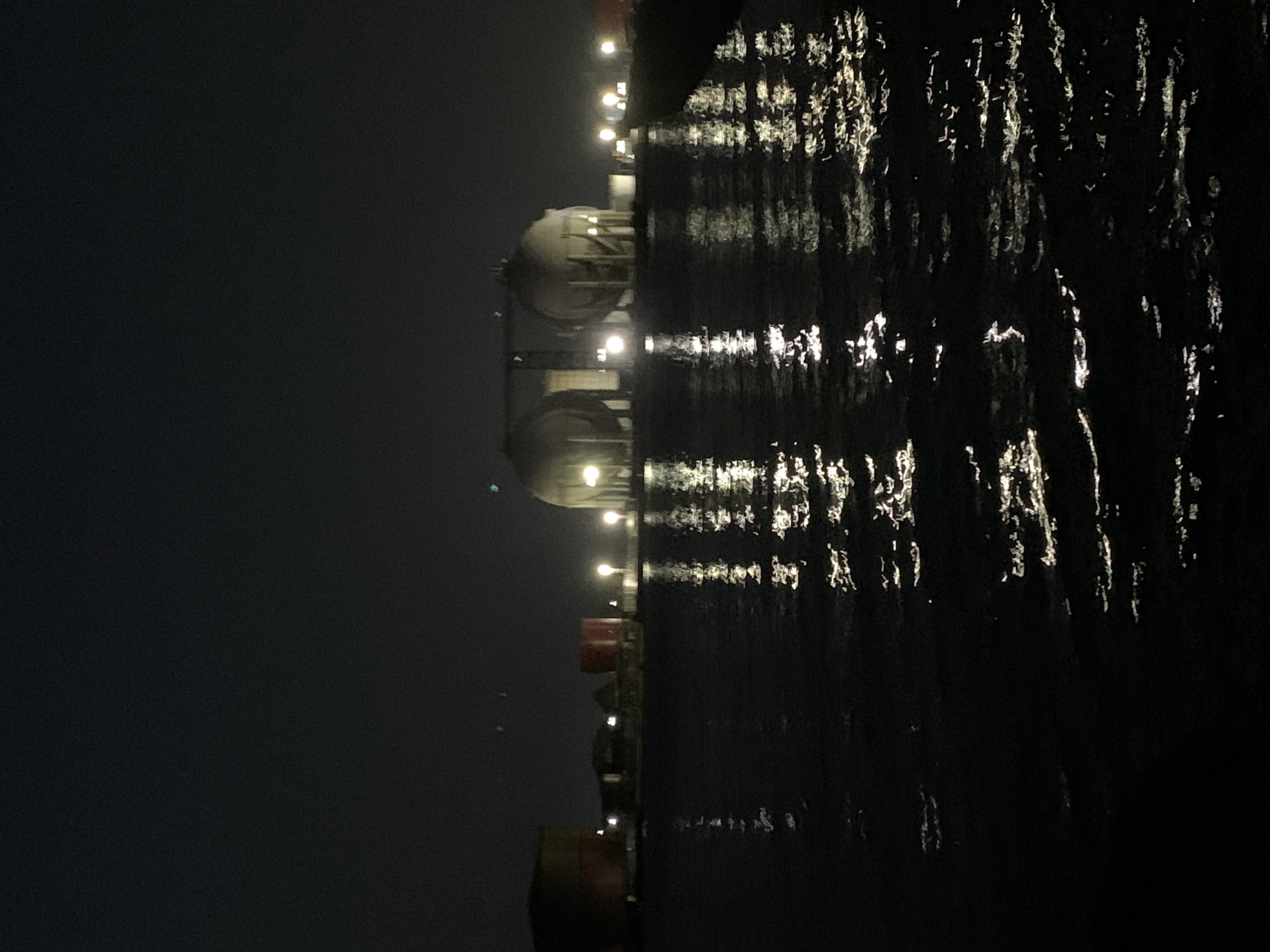 The image presents a nighttime scene of an industrial waterside environment, consistent with a location like Ibasa, Nigeria. The left side of the frame features prominent, large, light-colored structures, likely spherical or domed storage tanks, which are brightly illuminated by multiple artificial lights. These tanks appear to be part of a larger industrial facility or a large vessel, such as an LNG carrier, with visible structural supports. Below and between these tanks, other smaller, darker industrial components or parts of a dock are visible, also equipped with lights. Some reddish-brown elements are discernible in the lower left, possibly part of the vessel's hull or dock infrastructure.
The right side of the image is dominated by a dark body of water, which calmly reflects the numerous artificial lights. These reflections appear as elongated, shimmering vertical streaks, indicating a still or gently rippling water surface. The sky is a uniform, dark gray, suggesting a moonless night or heavy cloud cover, with artificial illumination being the primary light source. There are no people visible, and no specific activity is captured, presenting a static view of an industrial or maritime operational area after dark. The scene conveys a sense of industrial scale and quietness under the cover of night. No discernible text is visible in the image.